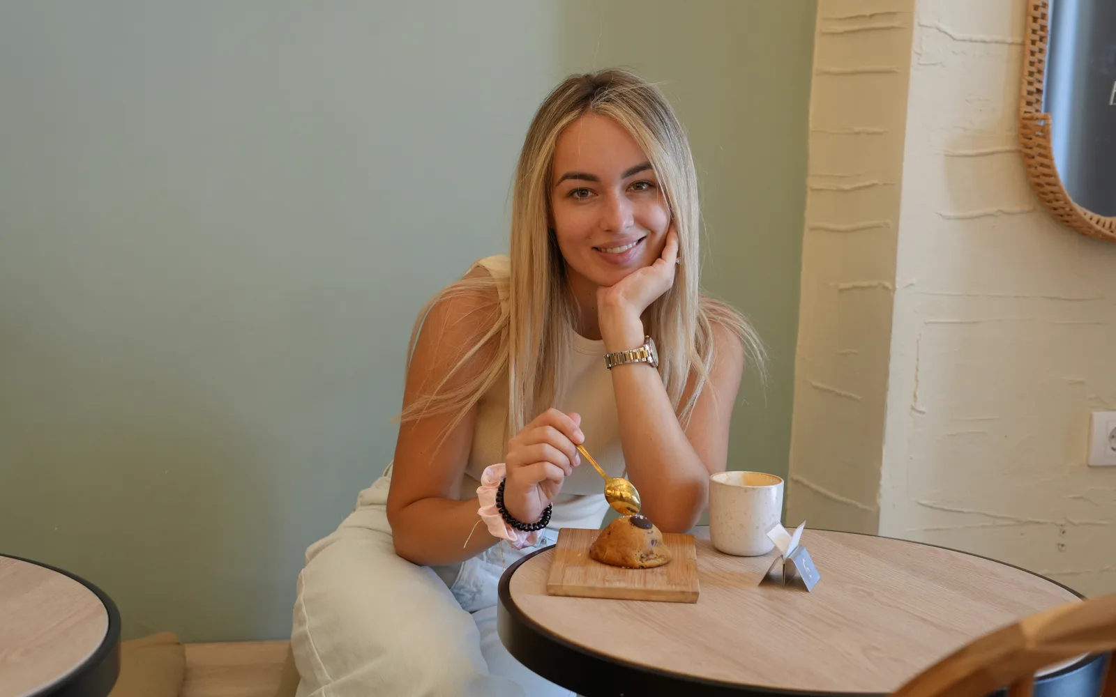 A guest at a round wooden table at Cookie Beacon Kralja Petra in Belgrade, spoon in hand over a chunky cookie on a board, with a cappuccino beside the sage-green wall and a raffia mirror