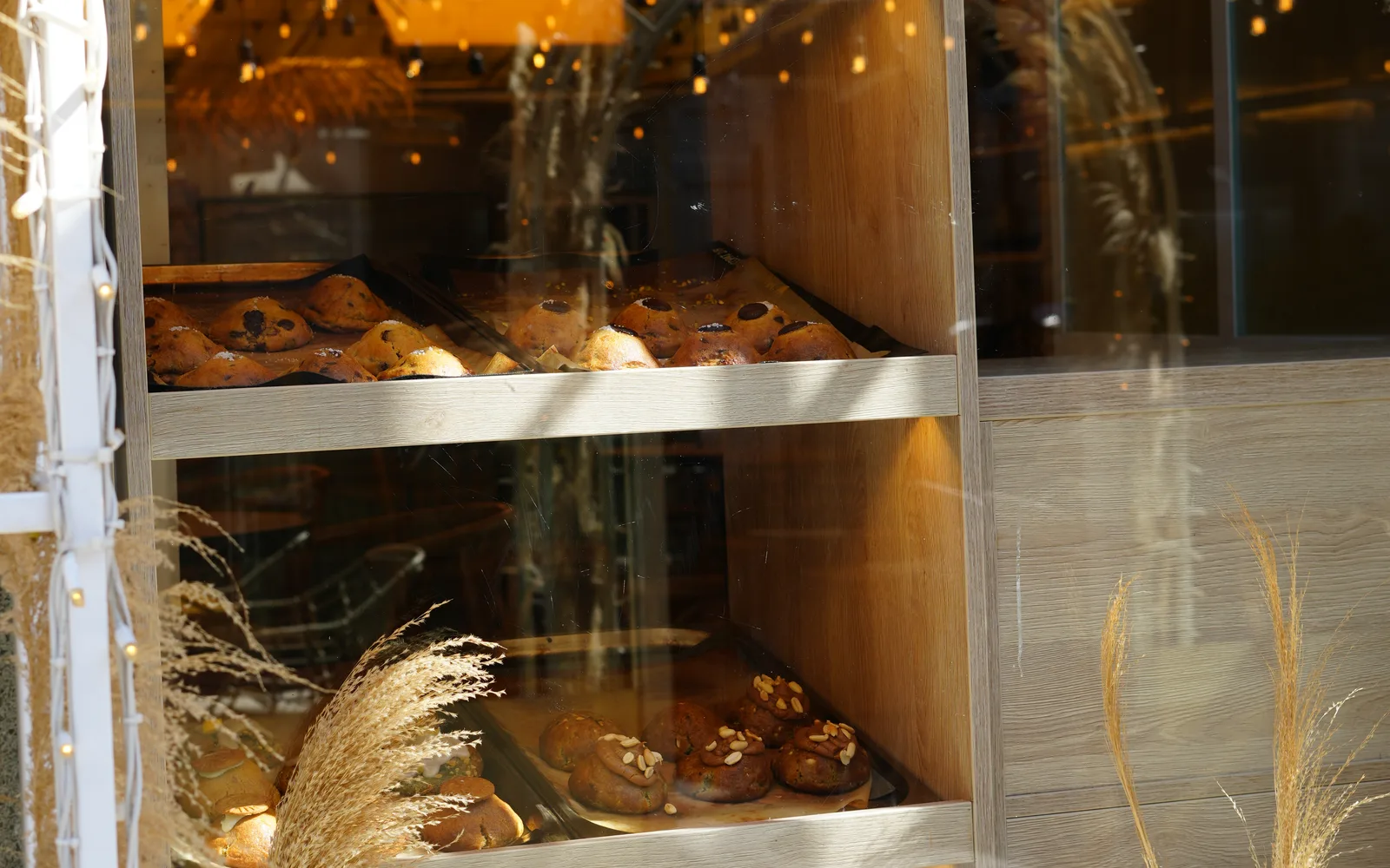 Trays of chunky cookies on lit wooden shelves behind the window at Cookie Beacon Kralja Petra in Belgrade, with pampas grass at the glass and the festoon lights reflected behind
