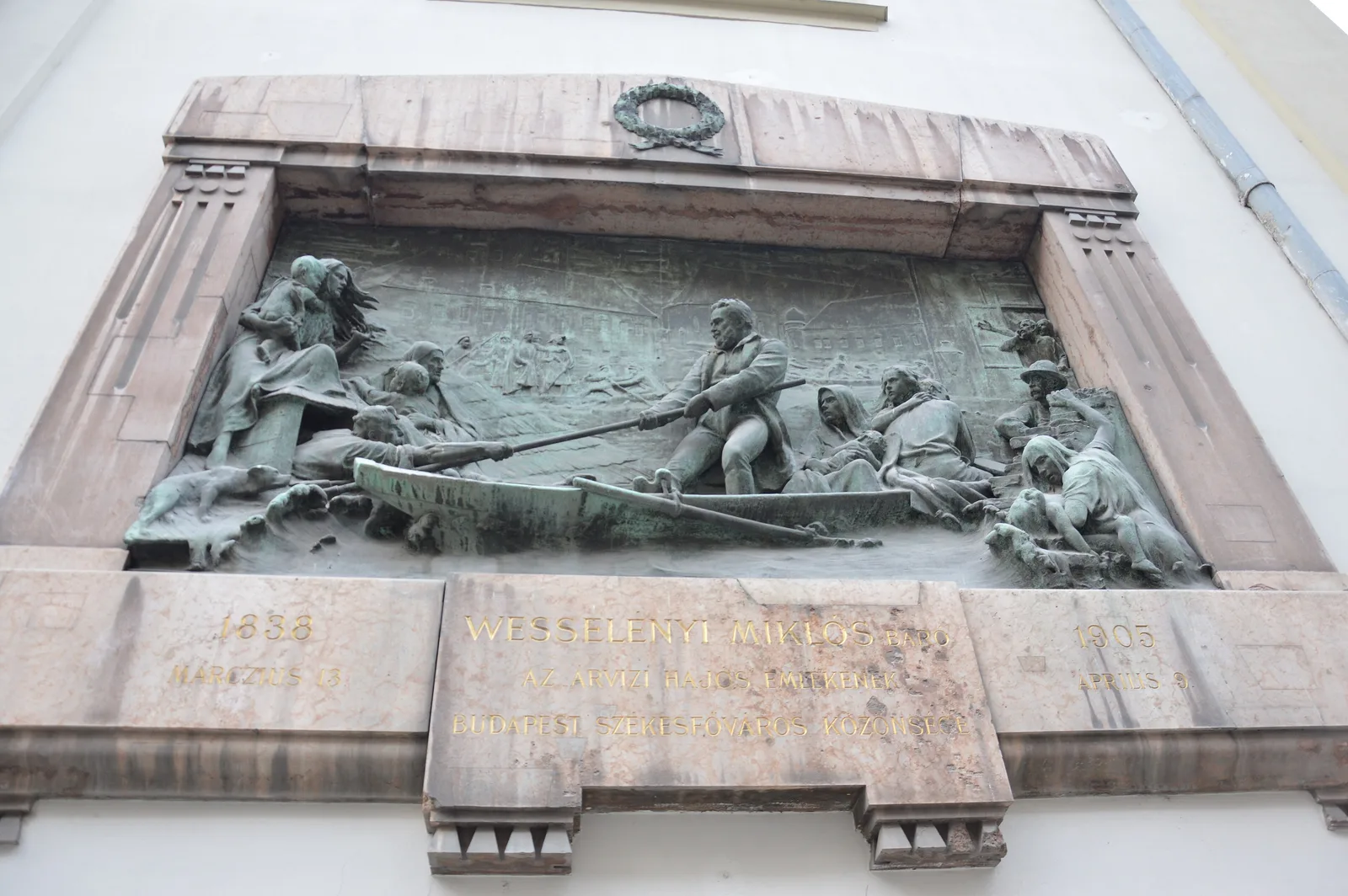 A bronze relief on the Franciscan Church in Budapest: a man in a rowboat holds out an oar to people on a rooftop in the 1838 flood, in a stone frame dated 1838 and 1905