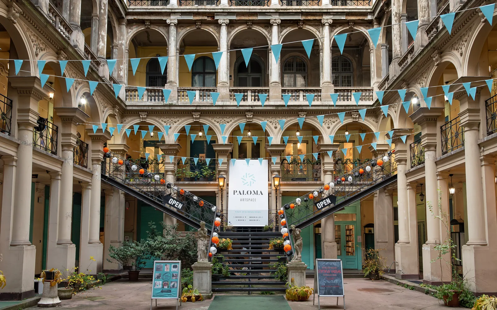 The Paloma courtyard in the Wagner house on Kossuth Lajos utca, Budapest: three floors of arcaded galleries around an iron staircase that splits into two arms, with a stone statue at each side of its foot