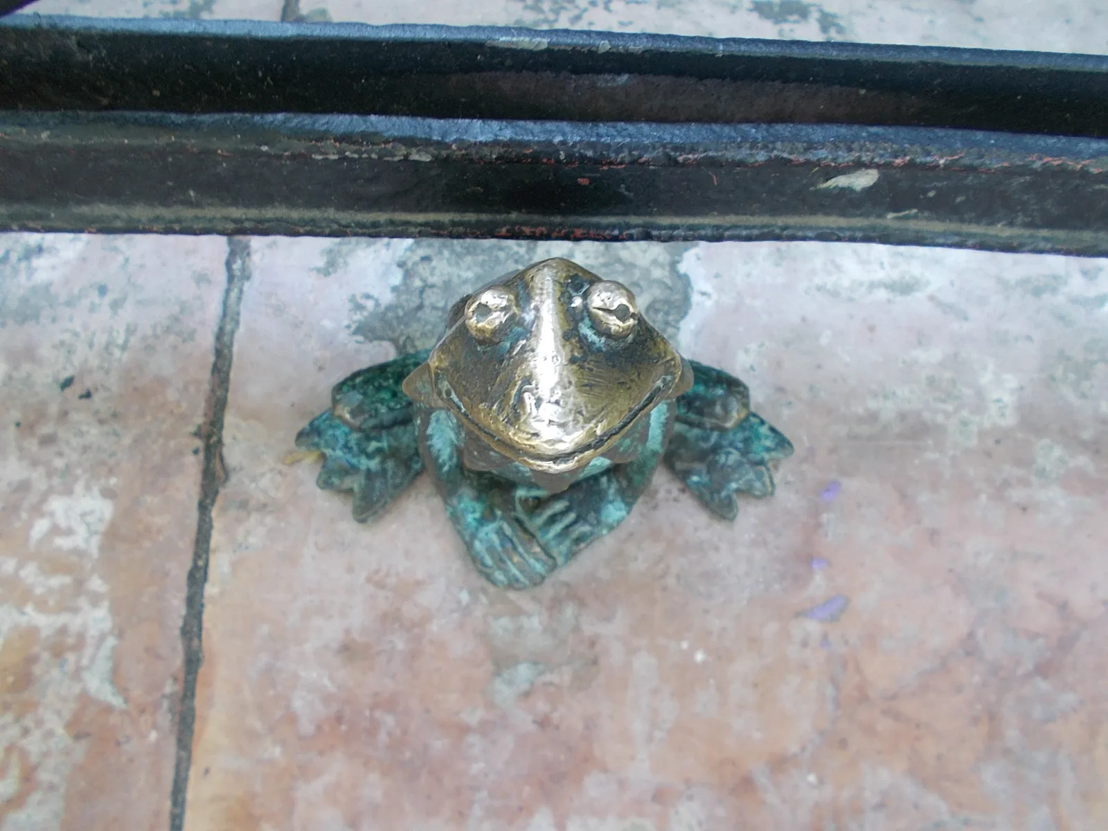 Mihály Kolodko's tiny bronze Kermit the Frog sitting on the paving stones of Szabadság tér in Budapest, under an iron fence rail, his nose rubbed shiny