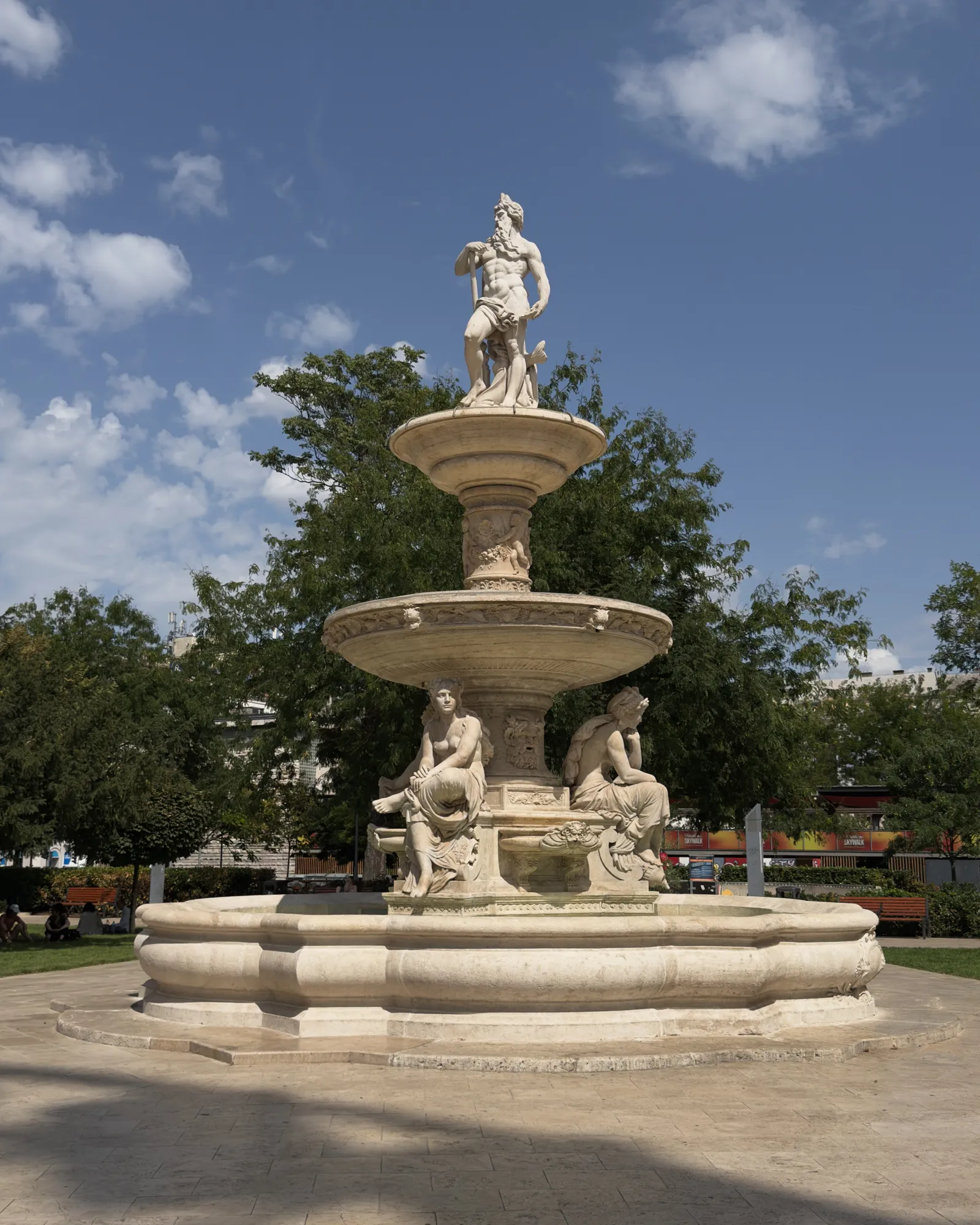 The Danubius Fountain on Erzsébet tér in Budapest: a pale stone fountain with a bearded river god standing on top and seated female figures around the lower basin, trees behind it