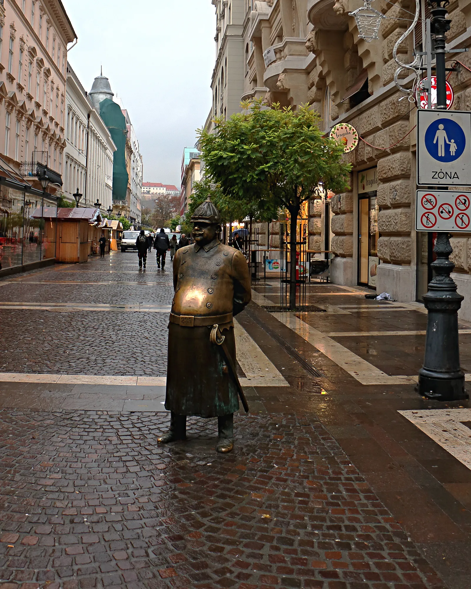 The Fat Policeman bronze statue on Zrínyi utca in Budapest, a round-bellied officer in a long coat and helmet standing on the paving stones of a pedestrian street, his belly polished bright