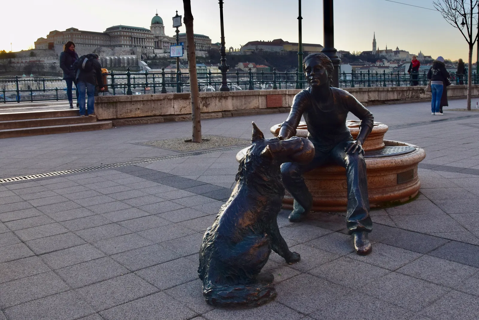 The Girl with Her Dog bronze statue by Dávid Raffay on Vigadó tér in Budapest: a young woman in jeans sitting on a round stone bench with her hand on a large bronze dog's head, Buda Castle across the Danube behind