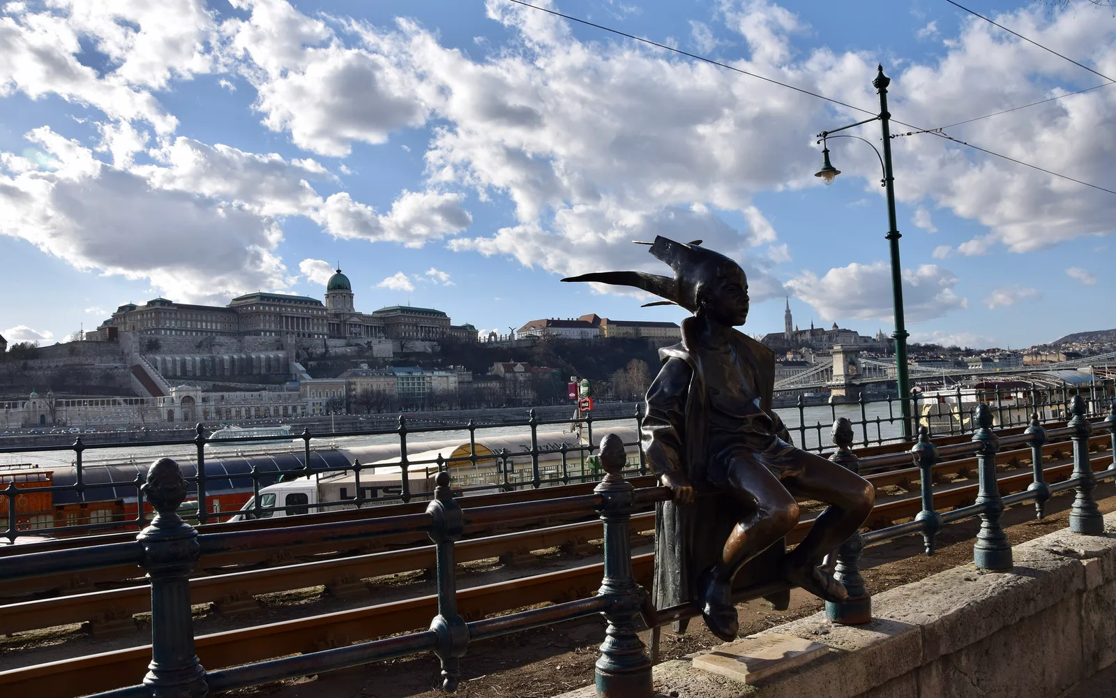 The Little Princess bronze statue in a paper crown and long coat, sitting on the iron railing of the Danube promenade in Budapest above the tram tracks, with Buda Castle across the river