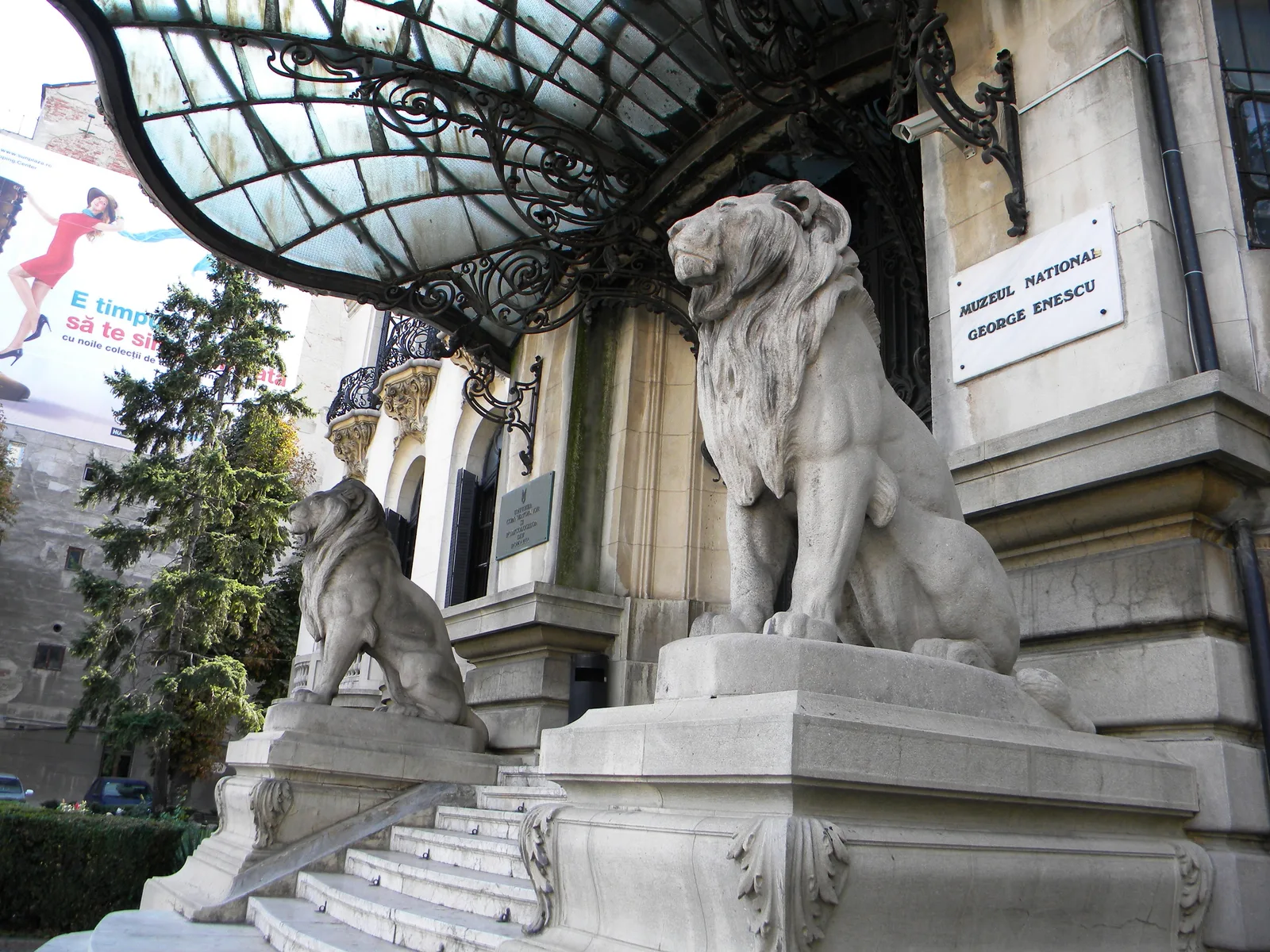 Two carved stone lions guarding the entrance of the Cantacuzino Palace at Calea Victoriei 141, Bucharest, under an iron-and-glass canopy, with the George Enescu museum sign by the door