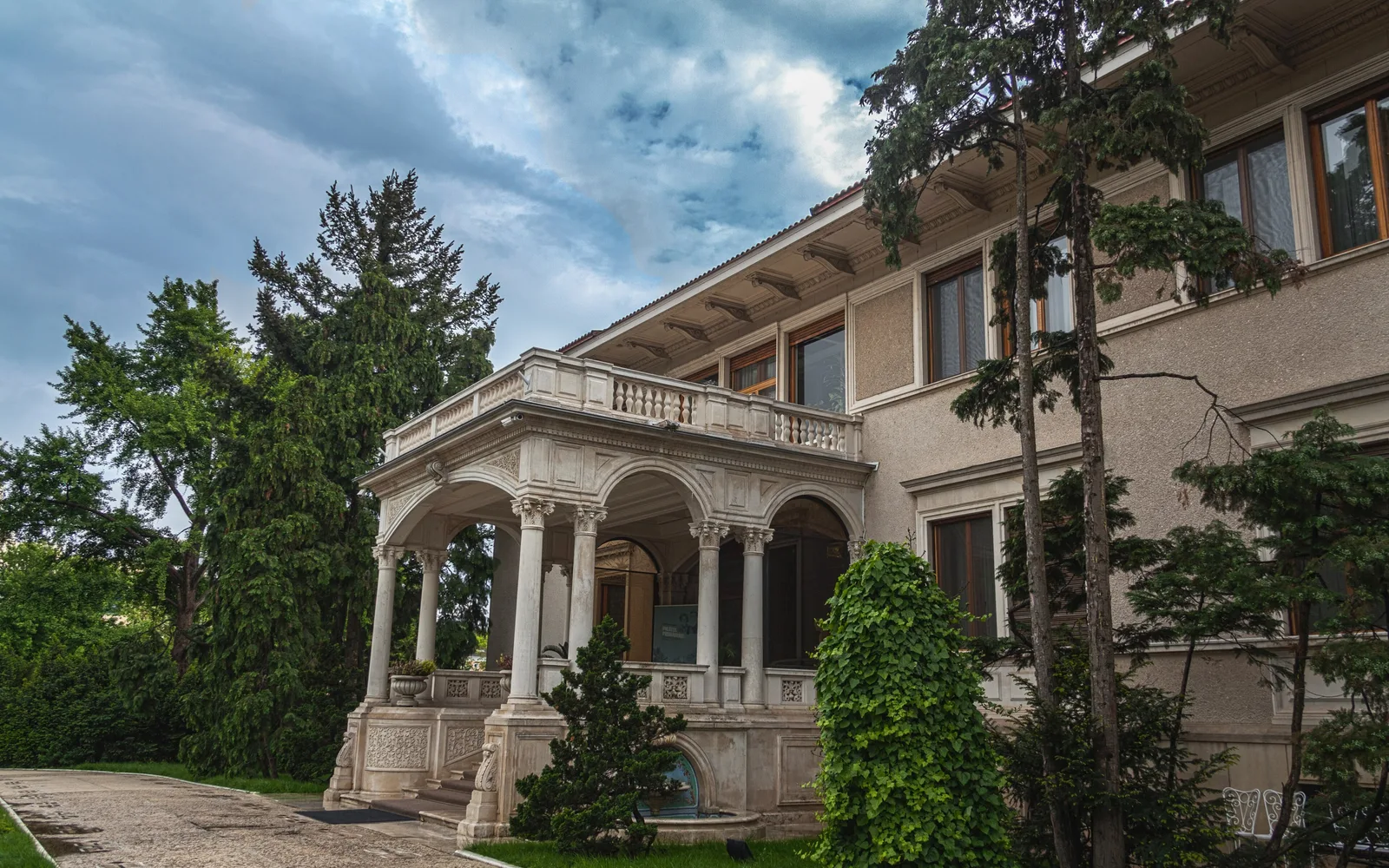 Intrarea în Palatul Primăverii din București: porticul cu arcade și coloane albe, cu terasă cu balustradă deasupra, între conifere