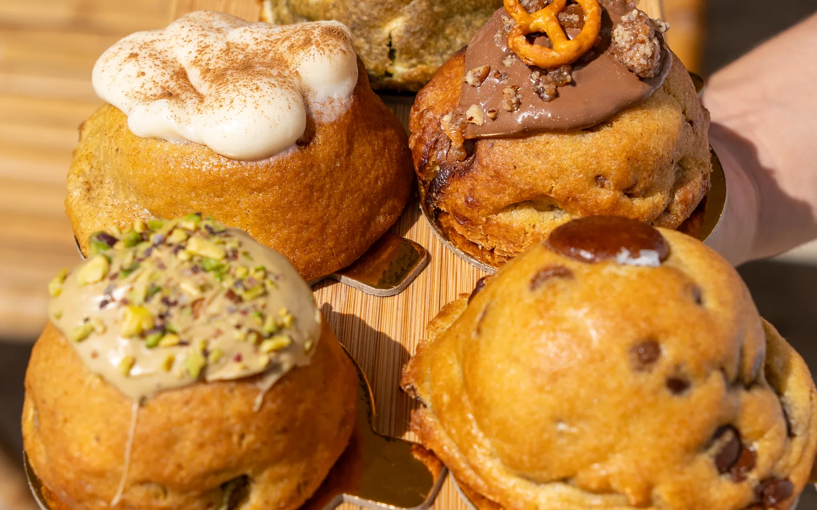 Handmade chunky cookies on a wooden board at Cookie Beacon, one topped with pistachio cream and crumbs, one with a pretzel, one with a cinnamon-dusted cream and a classic chocolate chip