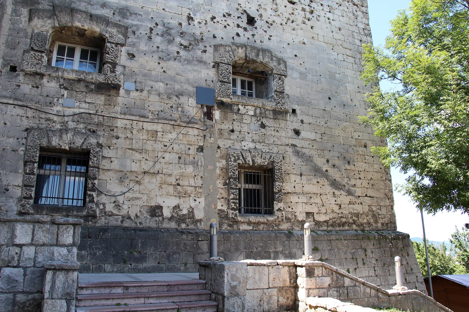 A stone wall of the Citadella on Gellért Hill, Budapest, covered in bullet and shell marks, with barred windows and steps at its foot