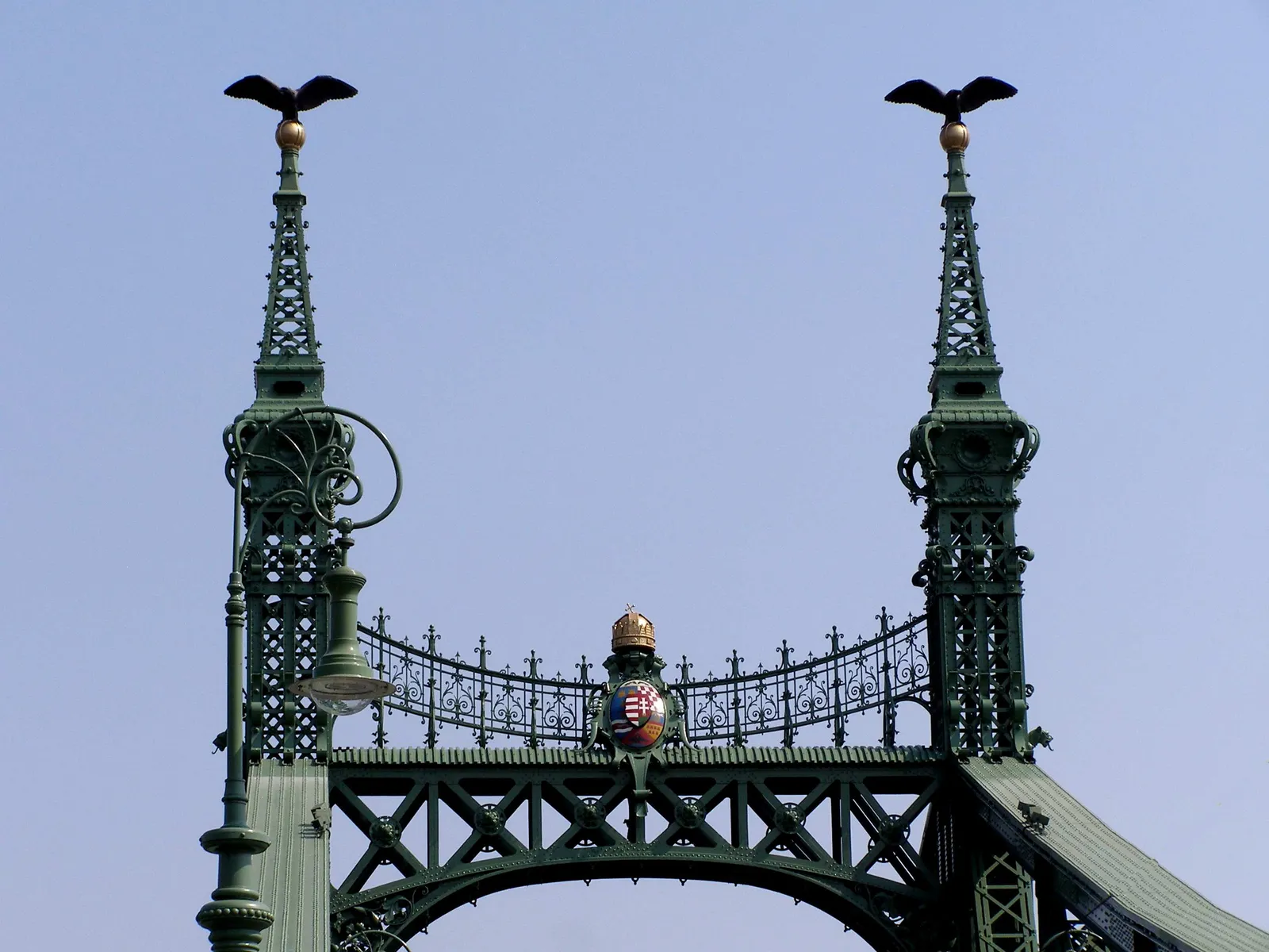 The top of Liberty Bridge in Budapest: two green iron masts each topped by a golden ball and a bronze turul bird, with the Hungarian coat of arms between them