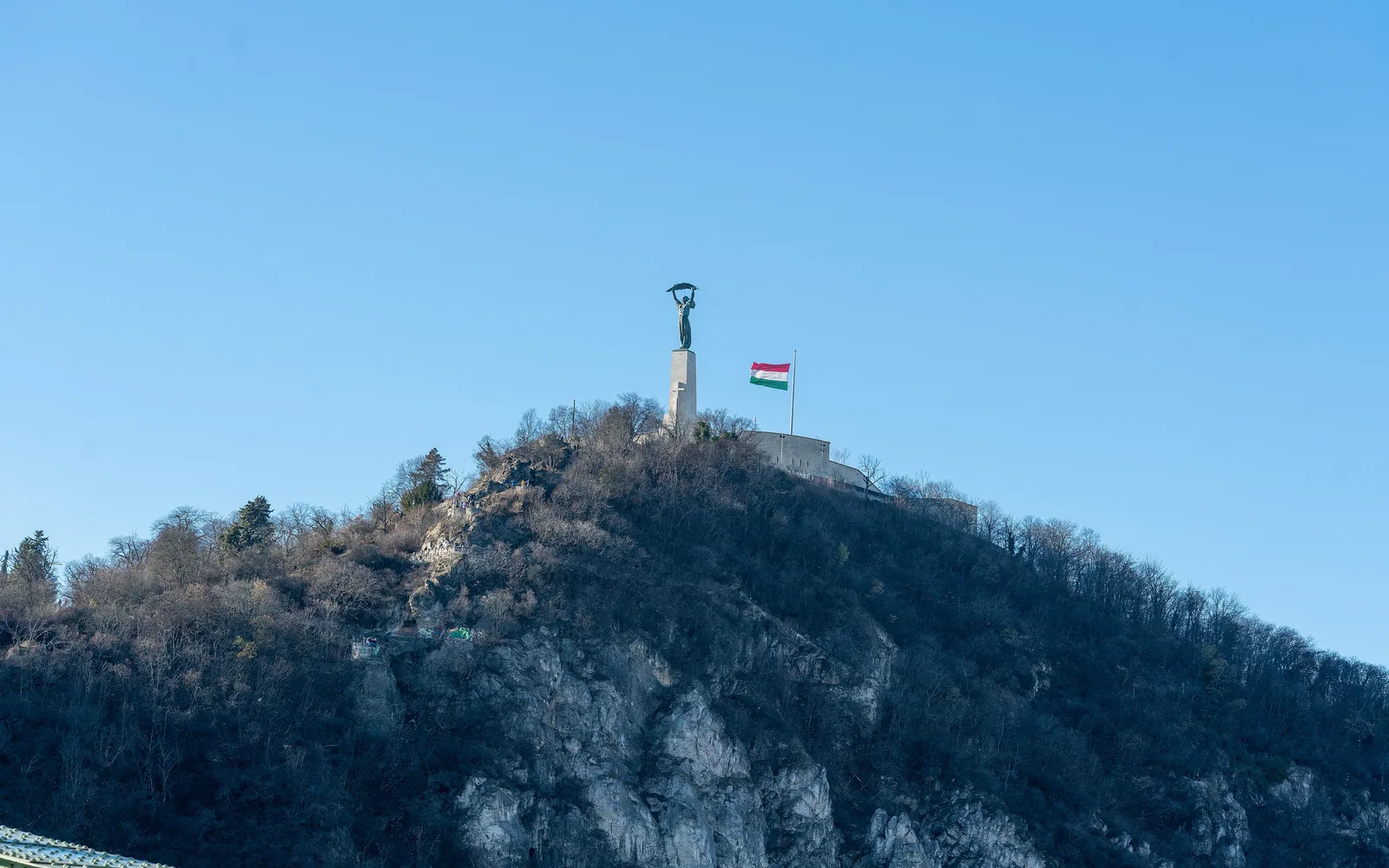 The summit of Gellért Hill in Budapest under a clear blue sky: the Liberty Statue holding a palm leaf above its stone pedestal, a Hungarian flag and the white walls of the Citadella behind the bare winter trees