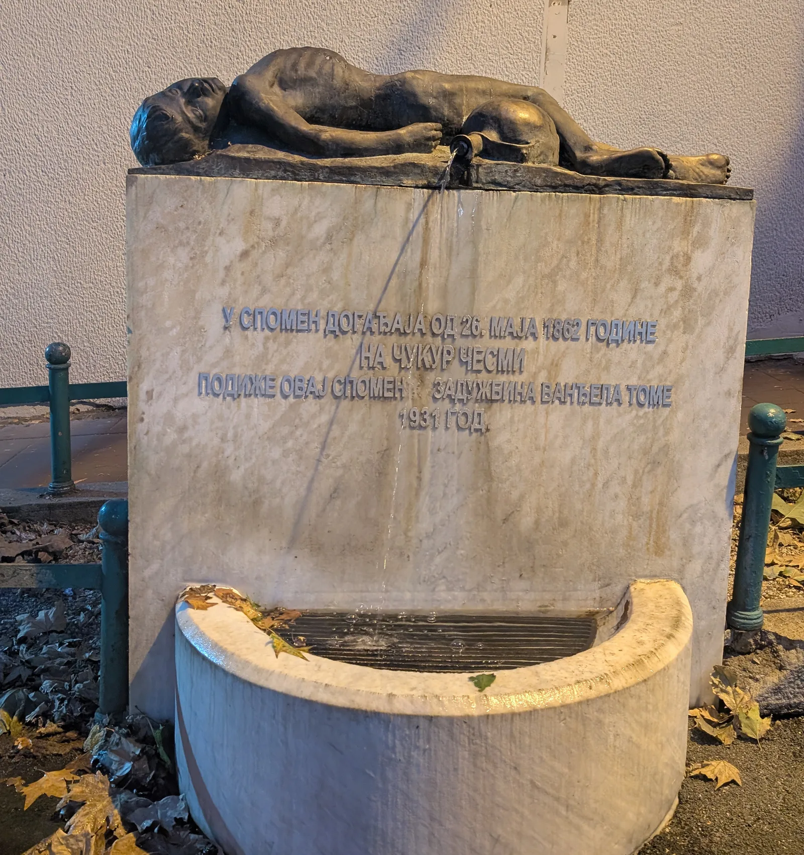 The Čukur fountain in Dorćol, Belgrade: a bronze boy lying on a marble block carved with a Cyrillic inscription dated 1862 and 1931, above a curved stone basin and autumn leaves