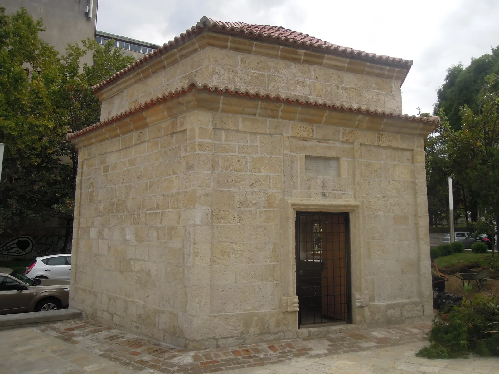 The small stone türbe of Sheikh Mustafa in Belgrade, with a tiled roof, a single barred door and a plain stone panel above it, on a paved corner between trees