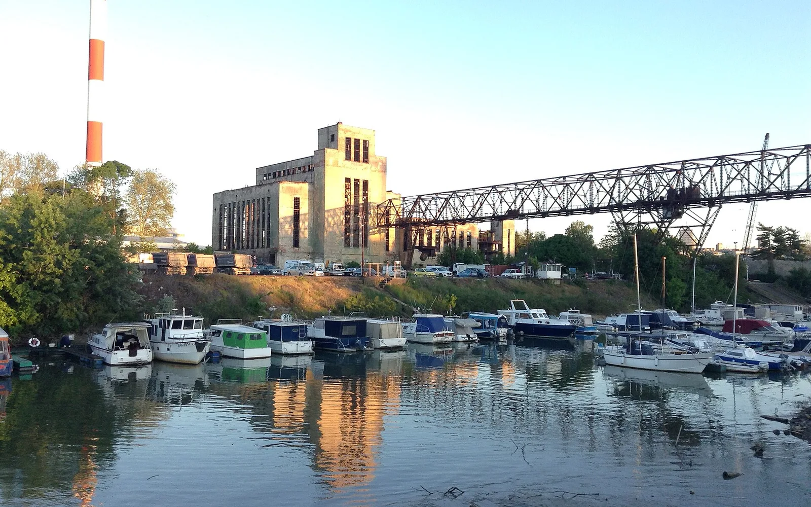 Small boats moored at Marina Dorćol on the Danube in Belgrade in warm evening light, with a long steel gantry and a tall industrial building behind the water