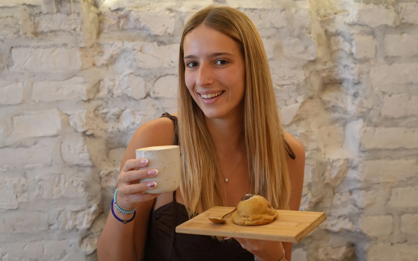 Gošća u Cookie Beacon-u na Kralja Petra drži šolju kafe i drvenu dasku sa chunky kolačićem, ispred zida od belih cigli