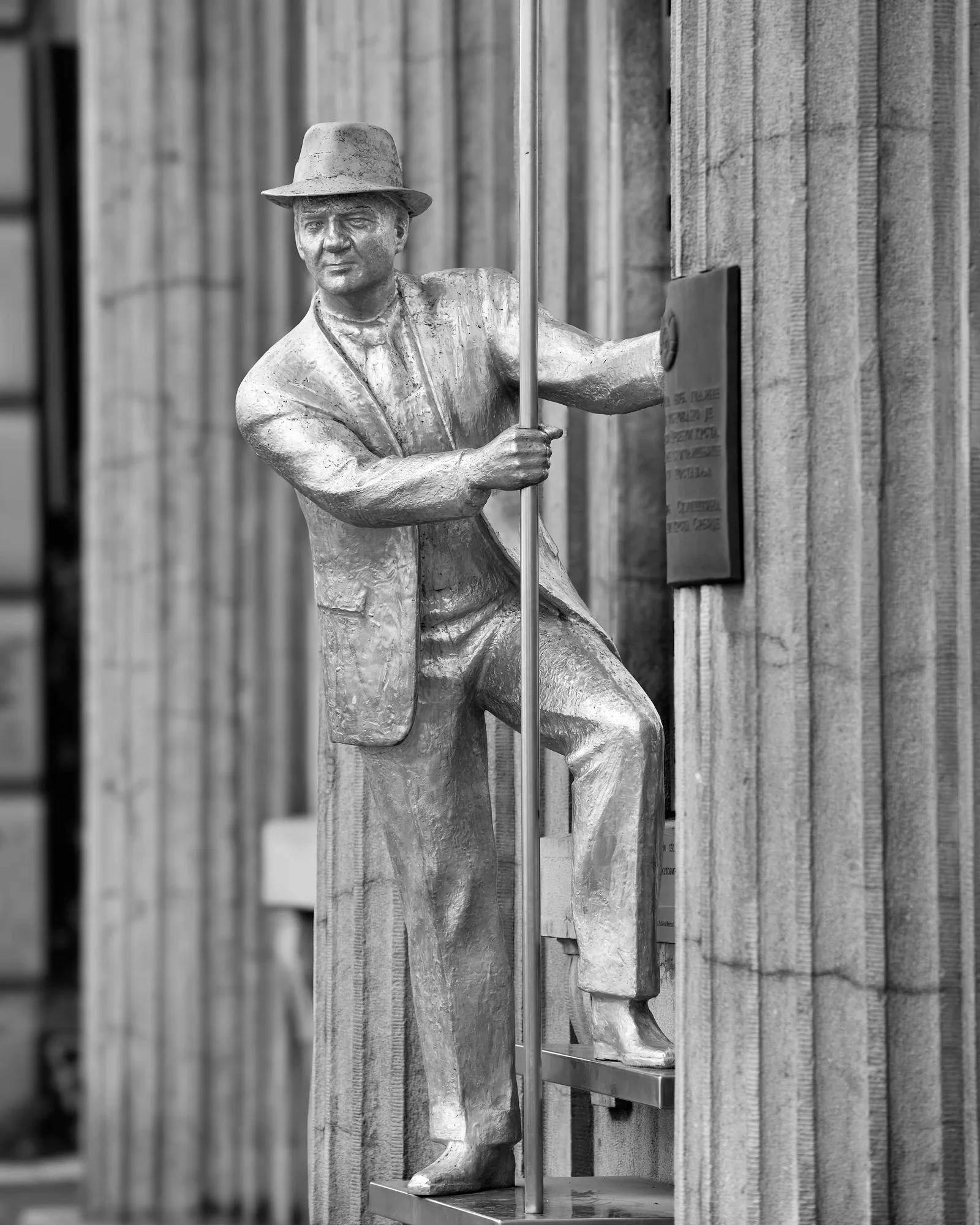 Black-and-white photo of the statue of actor Karl Malden in front of the Yugoslav Film Archive in Belgrade: a man in a hat and suit, one foot on a step, one hand gripping a tall pole beside a fluted stone column