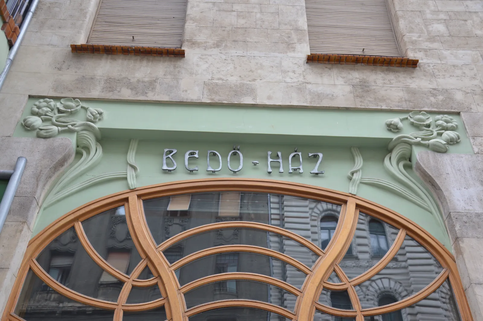 The metal letters "Bedő-Ház" on a pale green band with carved flowers, above a curved, honey-coloured wooden window frame on Bedő House, Honvéd utca, Budapest