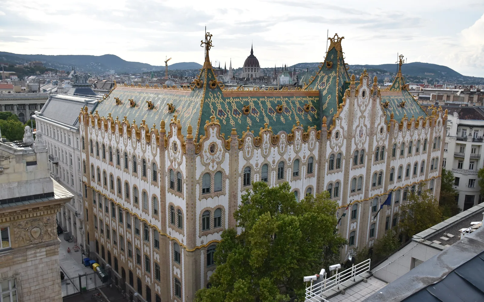 The former Postal Savings Bank on Hold utca, Budapest, seen from above: green Zsolnay roof tiles, gold ridges and the Parliament dome behind