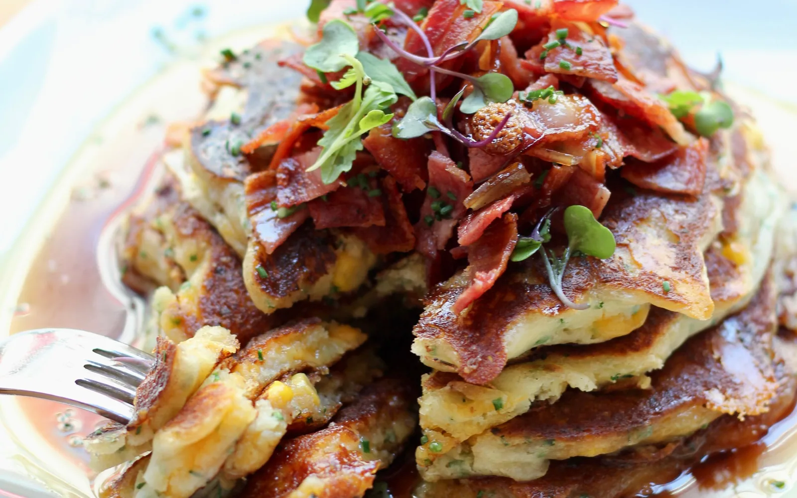 A stack of maple cheddar pancakes topped with crispy bacon and microgreens in maple syrup, a fork cutting into the corn and chive layers
