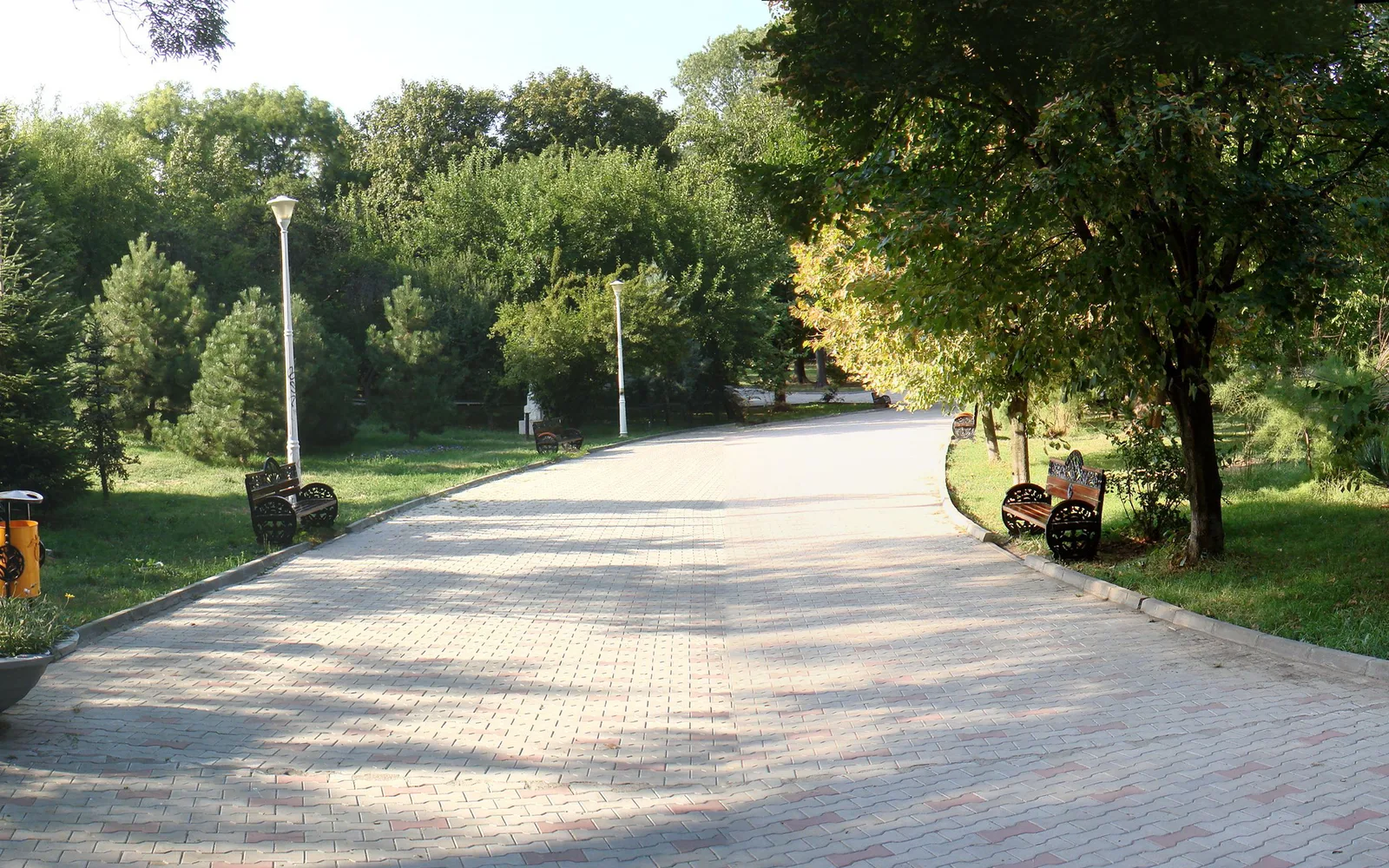 Alee pavată cu bănci de lemn cu laterale de fier și felinare albe, printre copaci, în Parcul Floreasca din nordul Bucureștiului