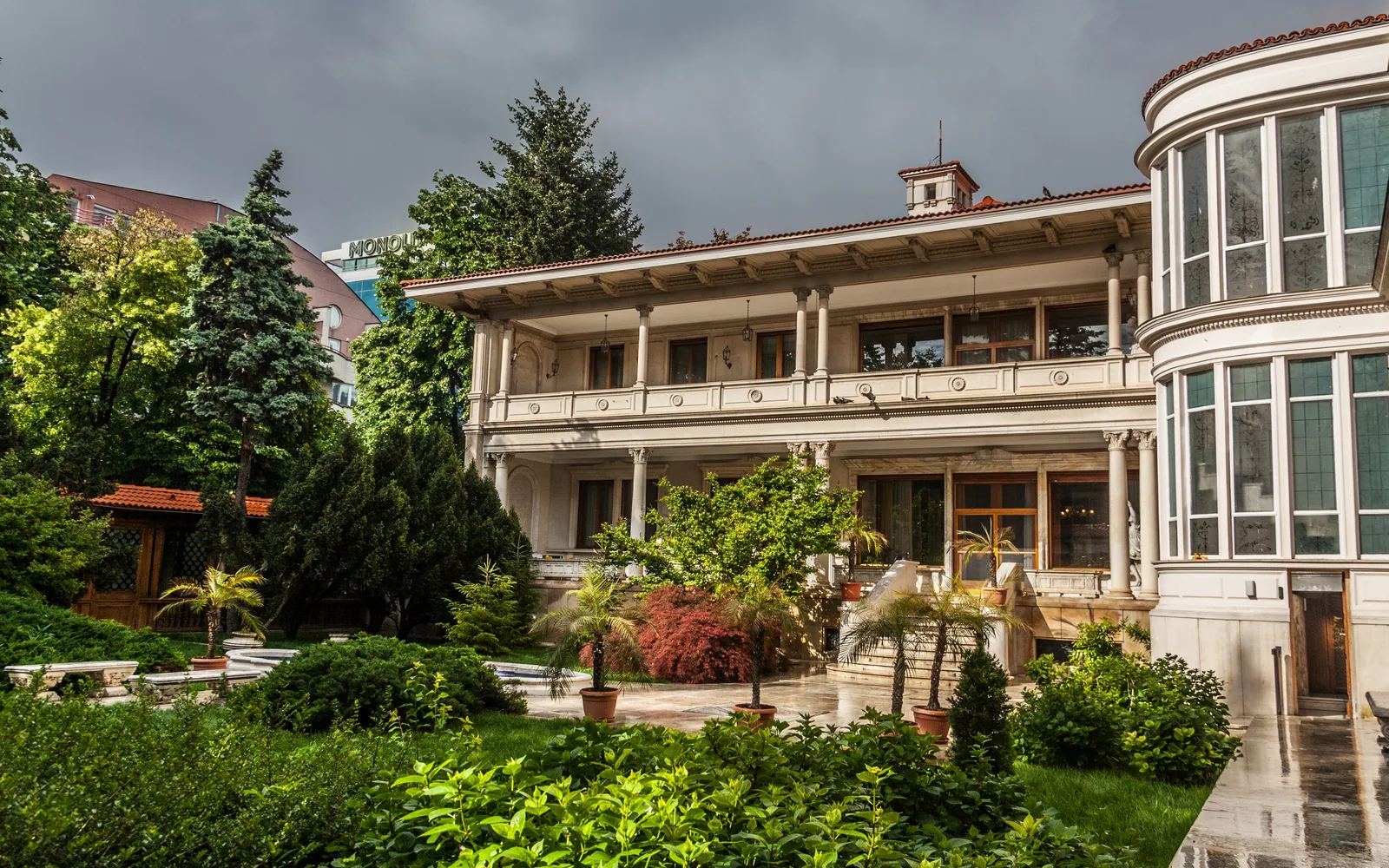 Garden front of the Primăverii Palace (Casa Ceaușescu) in Bucharest, a two-storey white villa with a columned loggia and a rounded glass corner, seen across clipped shrubs and potted palms