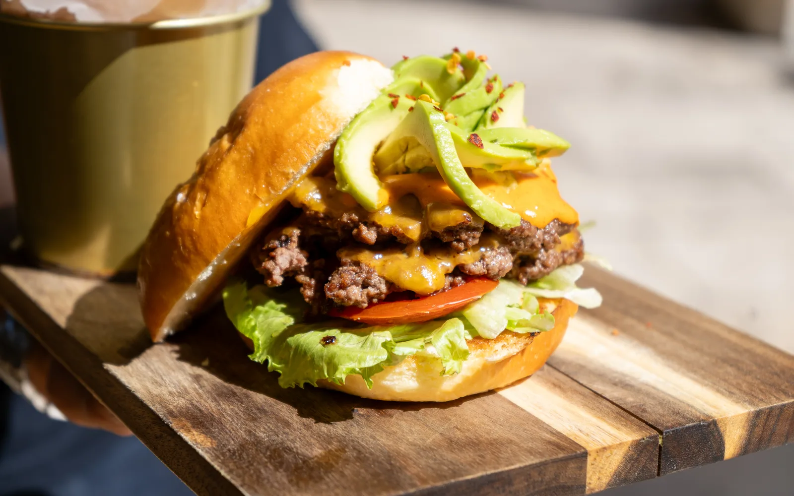 Close-up of the Avo Smashed Big Cheeseburger at Cookie Beacon Budapest: two smashed beef patties with crisp, craggy edges and melted cheddar, a slice of tomato and crunchy lettuce underneath, fanned avocado slices with chili flakes on top and the glossy pretzel bun lid tipped to one side