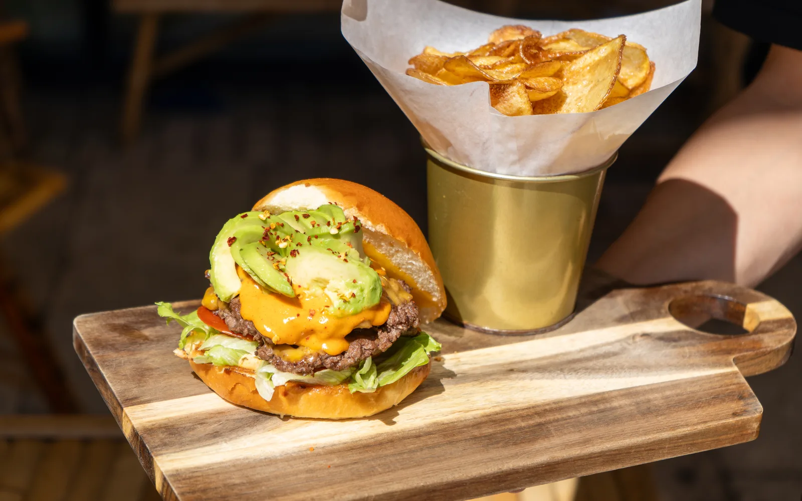 The Avo Smashed Big Cheeseburger carried out on its wooden board at Cookie Beacon Budapest: two smashed beef patties under melted cheddar and a glossy orange sauce, fanned avocado slices with chili flakes on top, crunchy lettuce and tomato in a golden pretzel bun, and a paper-lined gold cup of homemade potato chips alongside