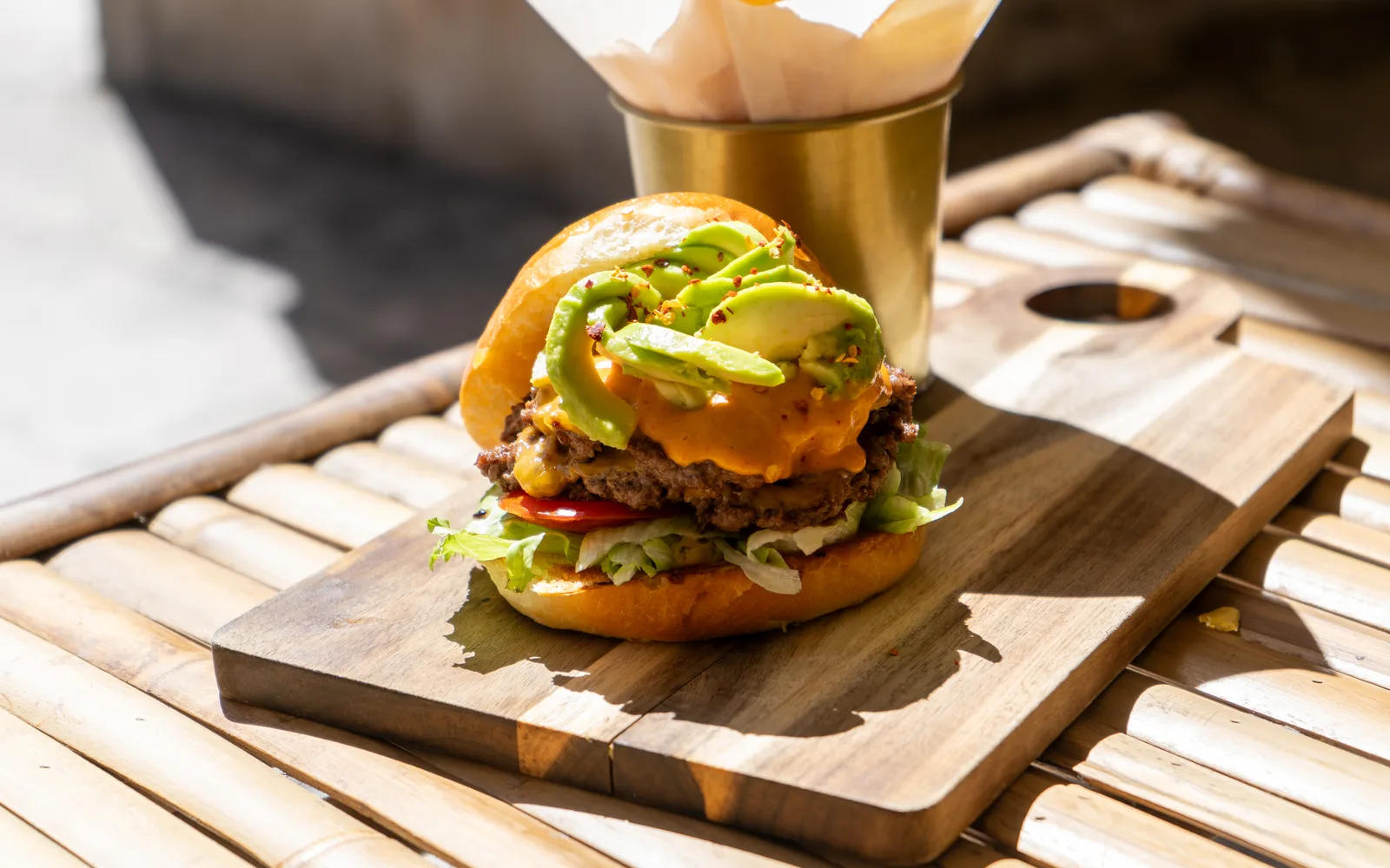 The Avo Smashed Big Cheeseburger on its wooden board on a sunny bamboo table at Cookie Beacon Budapest: fanned avocado slices and chili flakes over an orange sauce and a crisp-edged smashed patty, with tomato and lettuce in a golden pretzel bun and a paper-lined gold cup behind