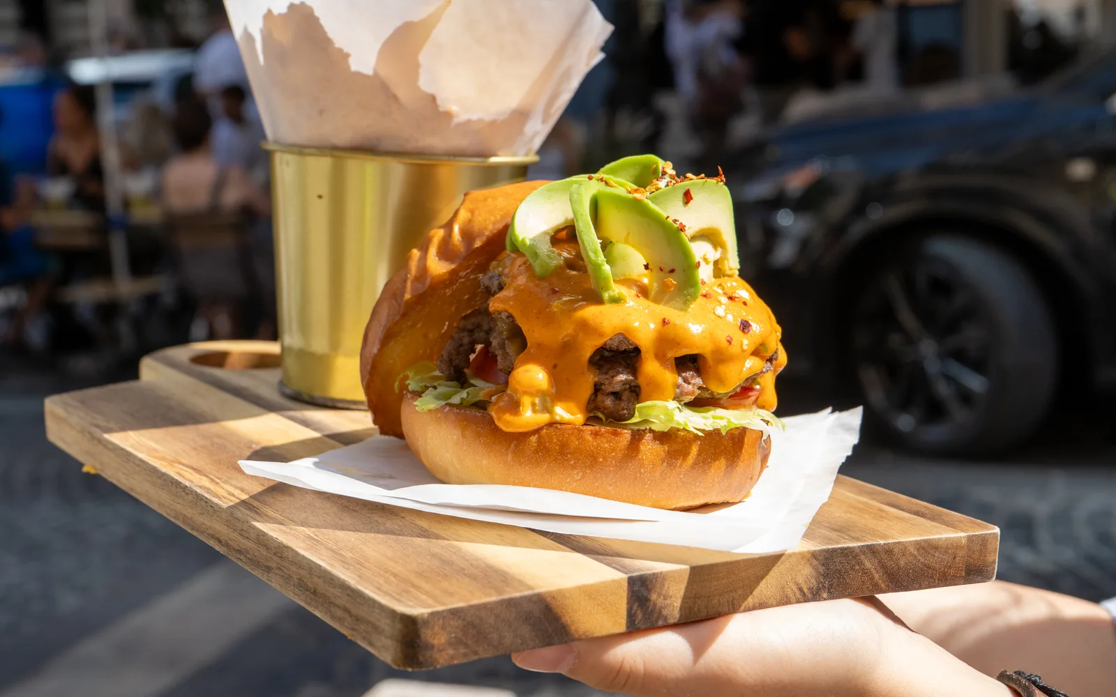 An Avo Smashed Big Cheeseburger from the Cookie Beacon Budapest menu on a wooden board: smashed beef patties, fanned avocado, melted cheddar sauce and chili flakes in a pretzel bun