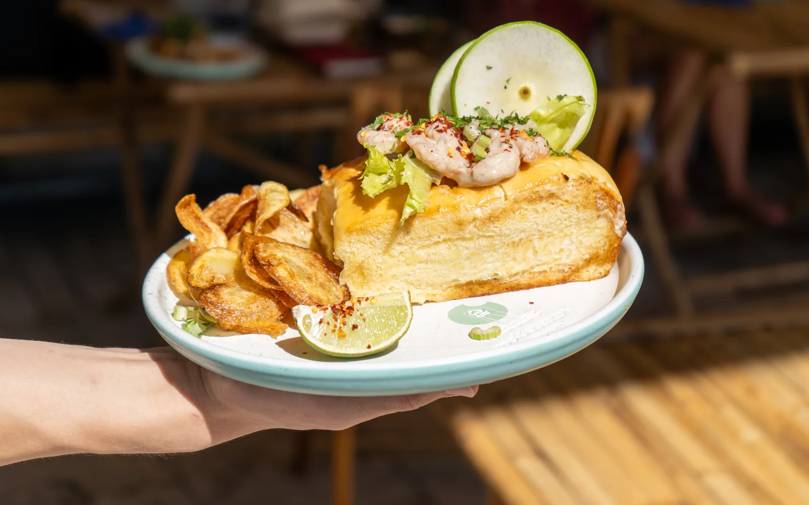 A Cape Cod Shrimp Roll carried across the terrace at Cookie Beacon Budapest: the toasted brioche roll with its shrimp topping, homemade chips and a citrus wedge on an aqua-rimmed plate, café tables blurred behind