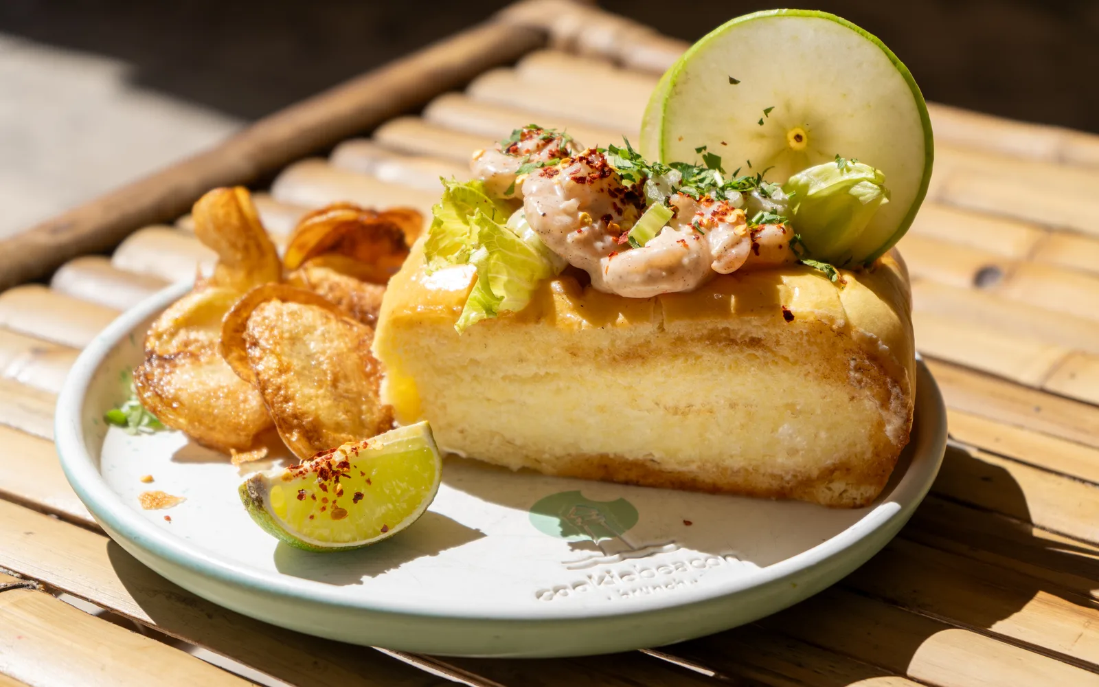 The Cape Cod Shrimp Roll on a sunny bamboo table at Cookie Beacon Budapest: a buttered, toasted brioche roll topped with shrimp in aioli, diced celery and chili flakes, with homemade chips alongside and a citrus wedge on the plate