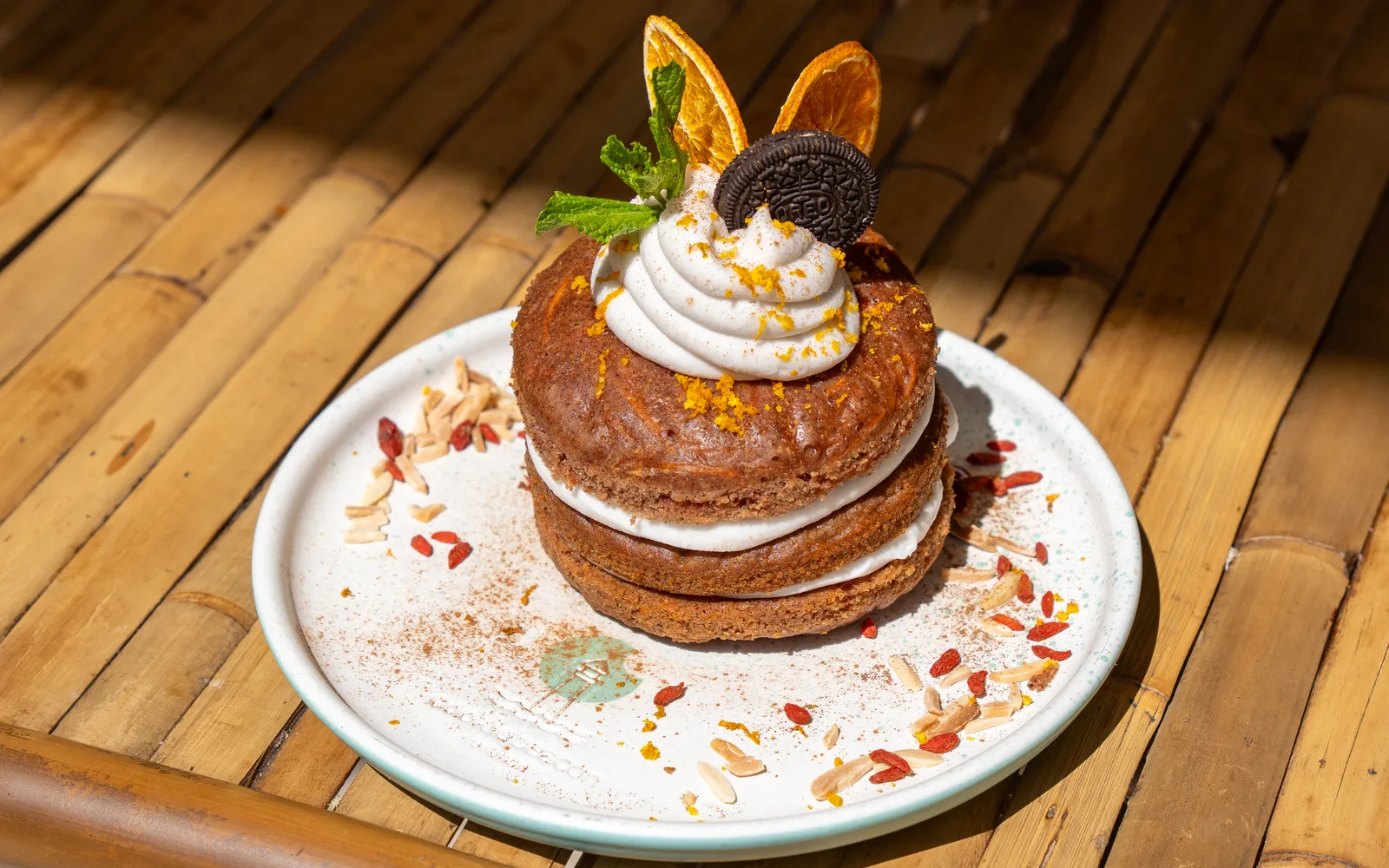 Carrot-Cake Pancakes on a bamboo table at Cookie Beacon Budapest: three thick pancakes layered with white cream, topped with a swirl of cream, orange zest, an Oreo, a sprig of mint and two dried orange slices, on a white plate scattered with slivered almonds, goji berries and cinnamon