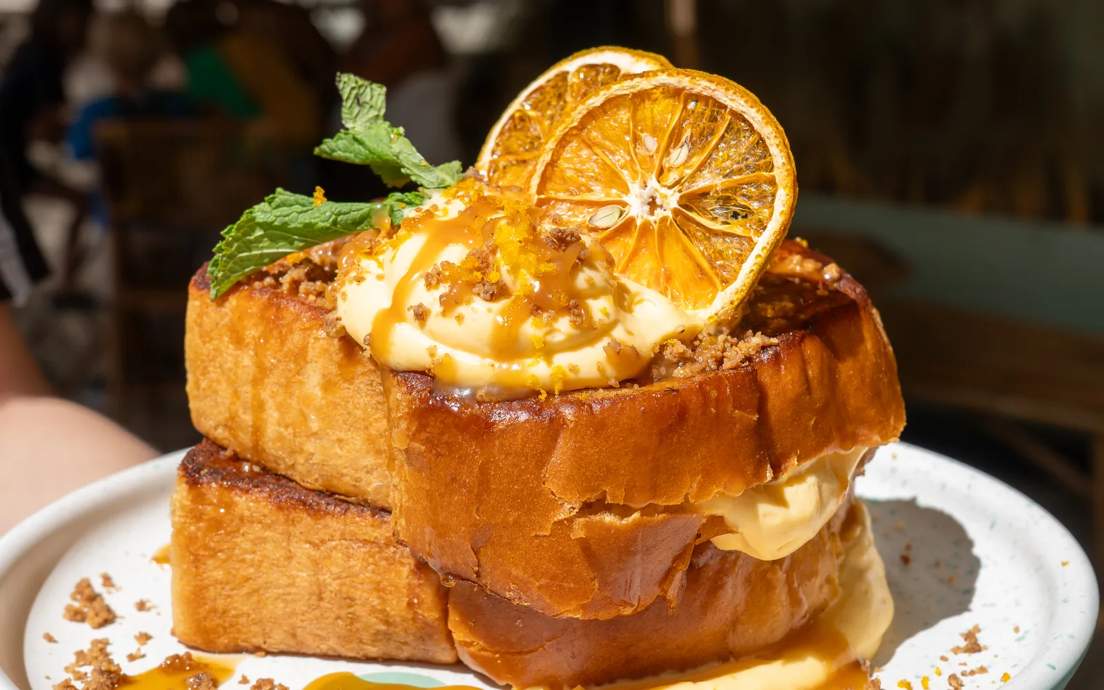 Crème Brûlée French Toast held out on a white plate at Cookie Beacon Budapest: two thick slices of golden brioche, one stacked slightly off-centre on the other, a swirl of orange pastry cream on top and squeezing out between the slices, cinnamon crumble, a drizzle of salted caramel, two dried orange slices and a sprig of mint