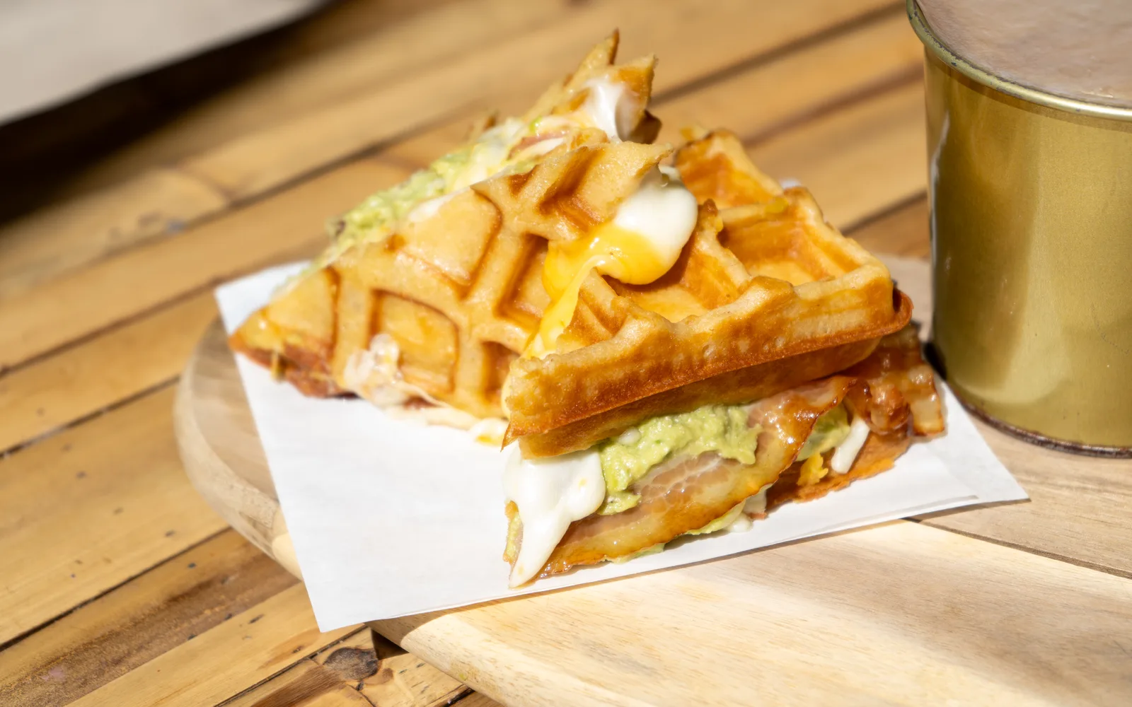 Close-up of the Grilled-Cheese Waffle-Sandwich at Cookie Beacon Budapest: melted cheese running from the waffle grid, with smashed avocado and crispy bacon showing at the edge, on a wooden board in the sun