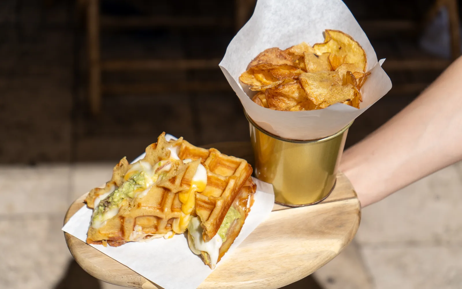 The Grilled-Cheese Waffle-Sandwich carried out on its wooden board at Cookie Beacon Budapest: two golden waffle halves on white paper with melted cheese and smashed avocado spilling from the sides, beside a paper-lined gold cup of homemade potato chips