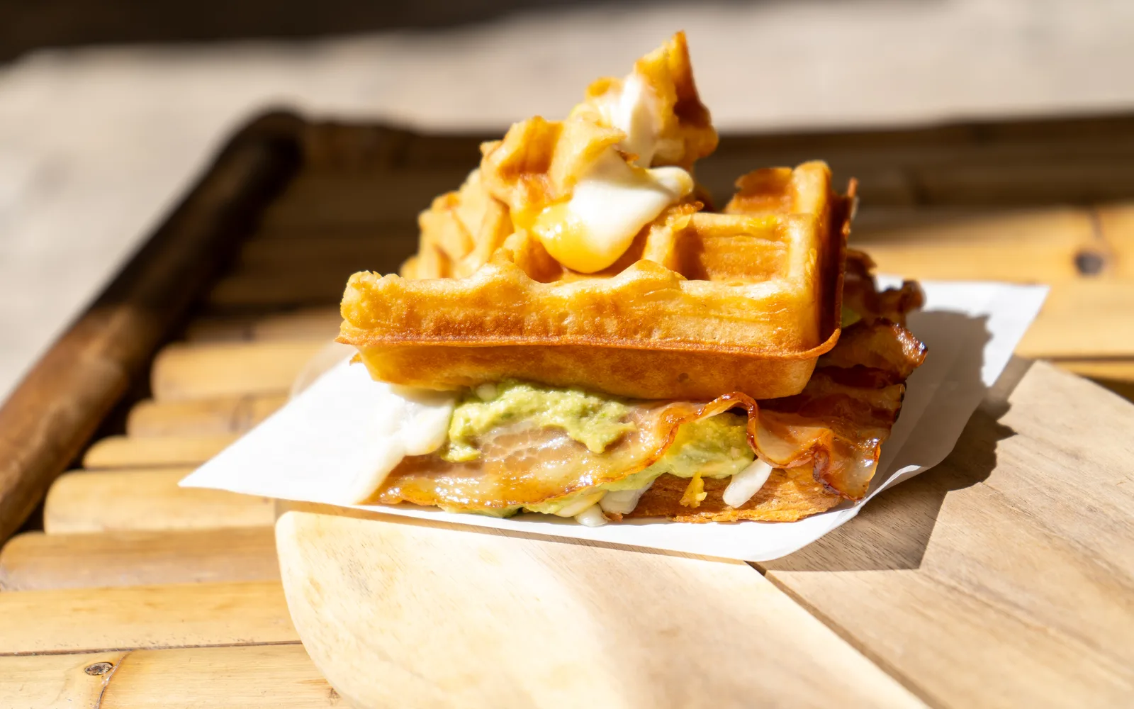 Side view of the Grilled-Cheese Waffle-Sandwich at Cookie Beacon Budapest: a crisp waffle lid over smashed avocado, crispy bacon and melted mozzarella, with the second half stacked on top, on white paper and a wooden board