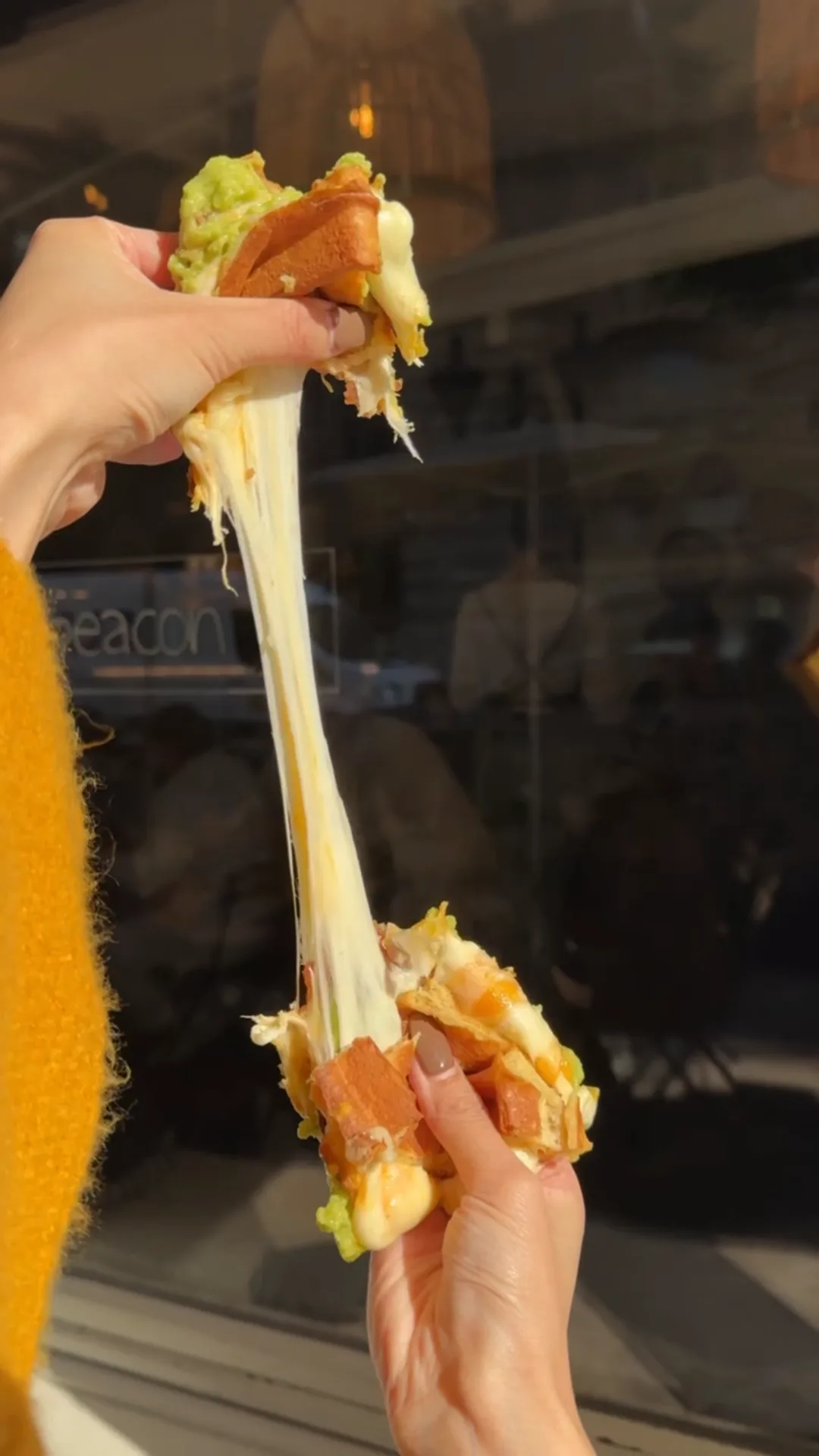 Two hands pulling a Grilled-Cheese Waffle-Sandwich apart in front of a Cookie Beacon window in Budapest, a long strand of melted cheese stretching between the two halves