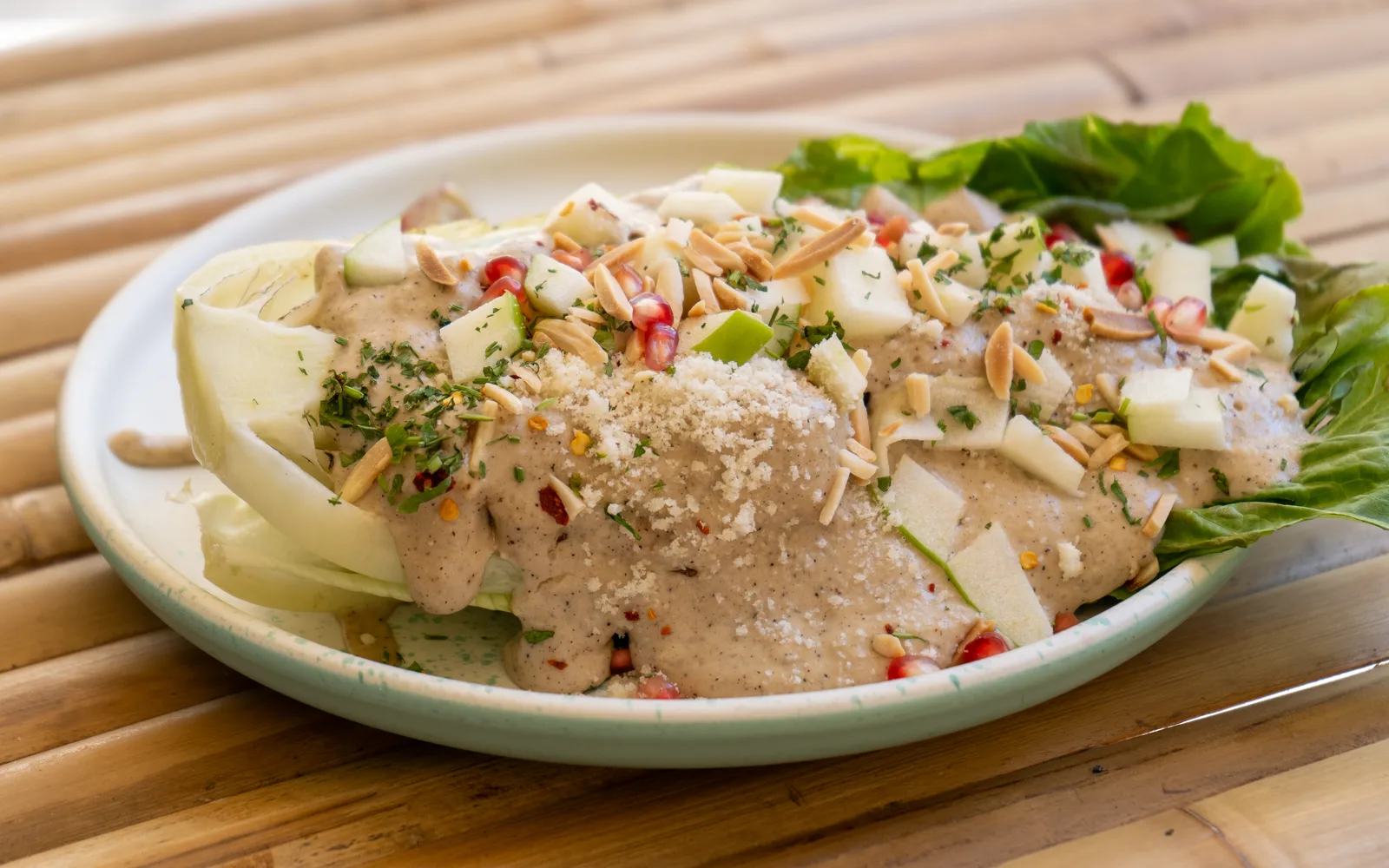 Close-up of the Grilled-Romaine Caesar Crunch on a bamboo table at Cookie Beacon Budapest: Caesar sauce poured over a halved head of romaine, topped with apple chunks, toasted almond sticks, pomegranate seeds and chopped green herbs