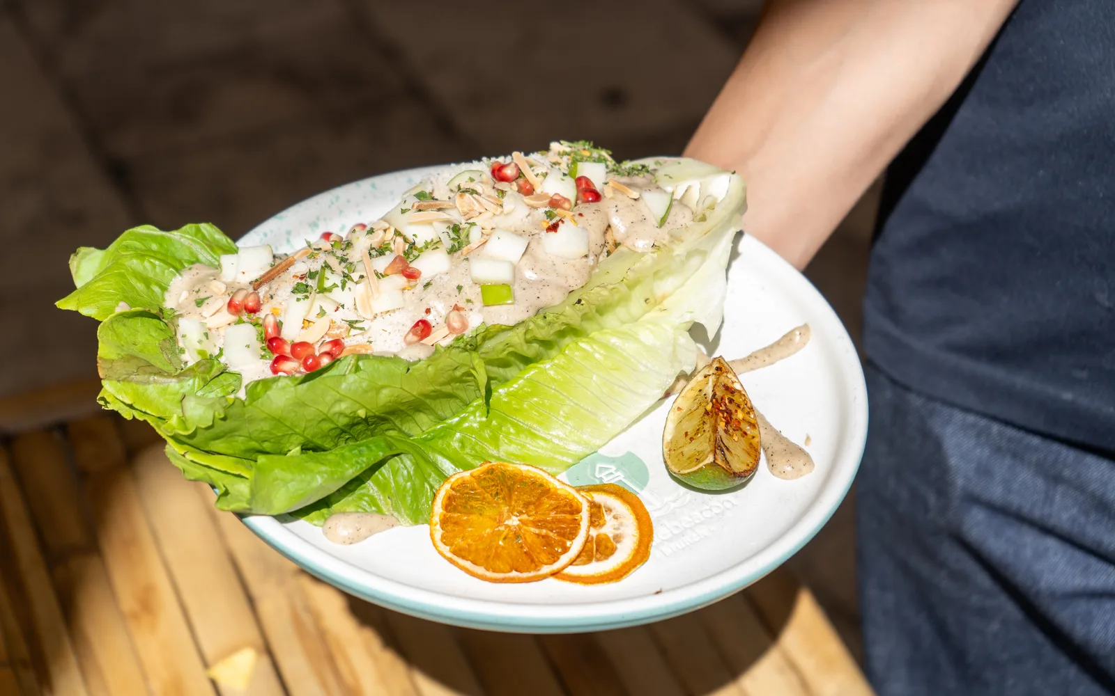 The Grilled-Romaine Caesar Crunch carried out on a white plate at Cookie Beacon Budapest: a halved head of romaine filled with pale Caesar sauce, white and green apple chunks, almond sticks, pomegranate seeds and chopped herbs, with dried orange slices and a charred citrus half on the rim