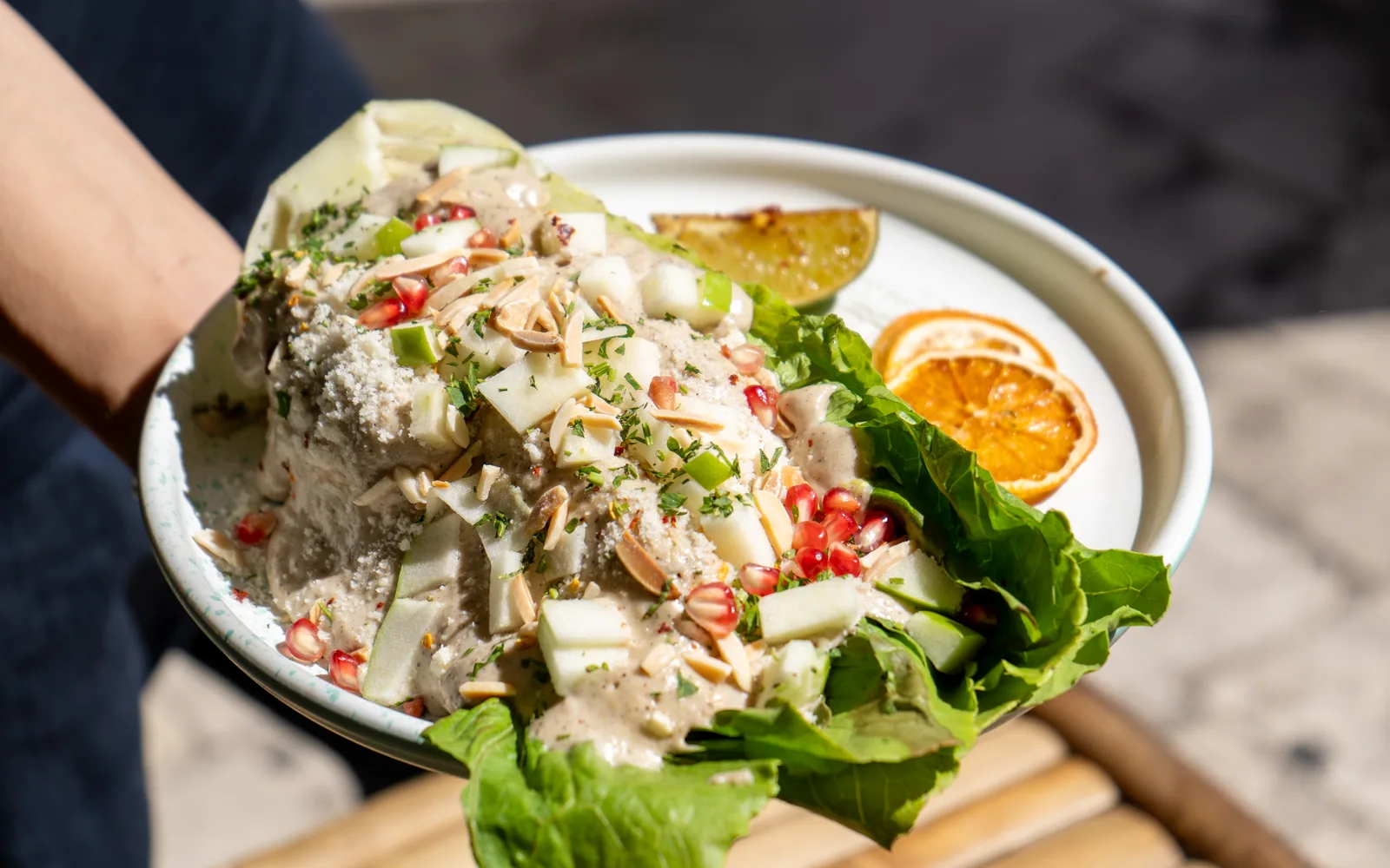 The Grilled-Romaine Caesar Crunch held out on a white plate at Cookie Beacon Budapest, seen from above: Caesar sauce over a halved head of romaine, covered with apple chunks, toasted almond sticks, pomegranate seeds and chopped herbs, the green leaf tips fanning over the rim, with dried orange slices and a charred citrus half behind