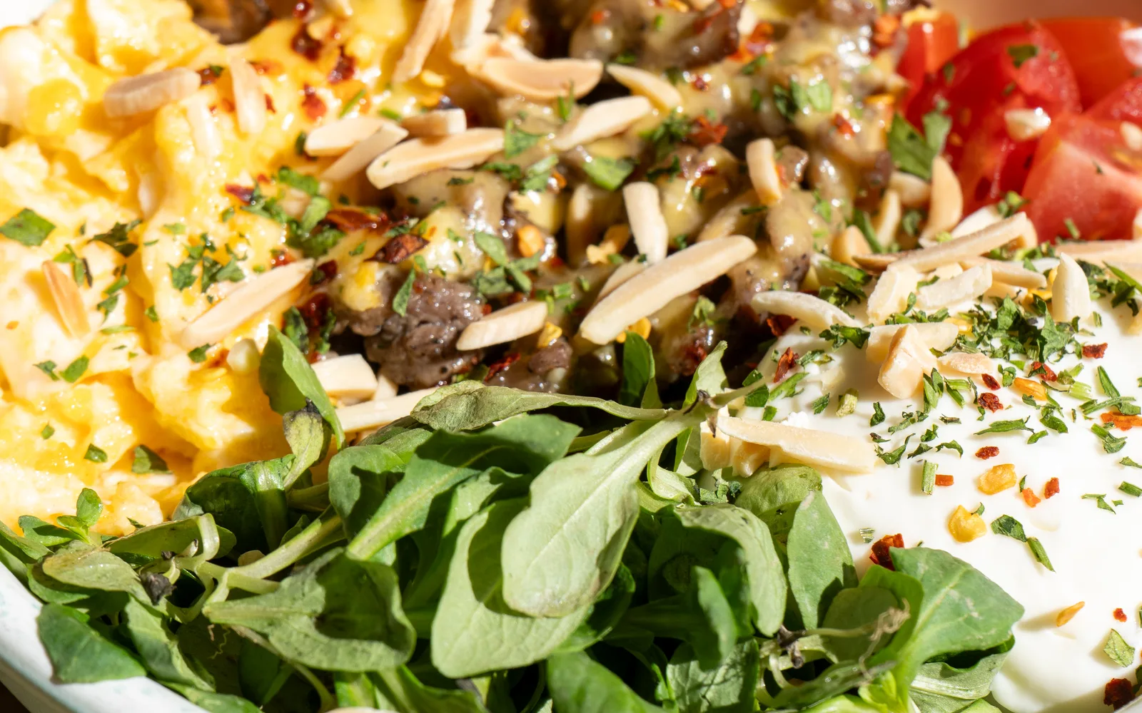Close-up of the Philly Cheese Steak Protein-Bowl at Cookie Beacon Budapest: sirloin in cheese sauce under slivered almonds and chili flakes, between scrambled eggs, cherry tomatoes, homemade sour cream and lamb's lettuce