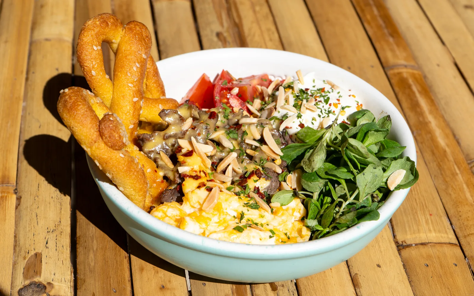The Philly Cheese Steak Protein-Bowl on a sunlit bamboo table at Cookie Beacon Budapest: scrambled eggs at the front, sirloin in cheese sauce in the middle, two sesame keto pretzels leaning on the rim, with lamb's lettuce, cherry tomatoes and homemade sour cream, all scattered with slivered almonds and chili flakes