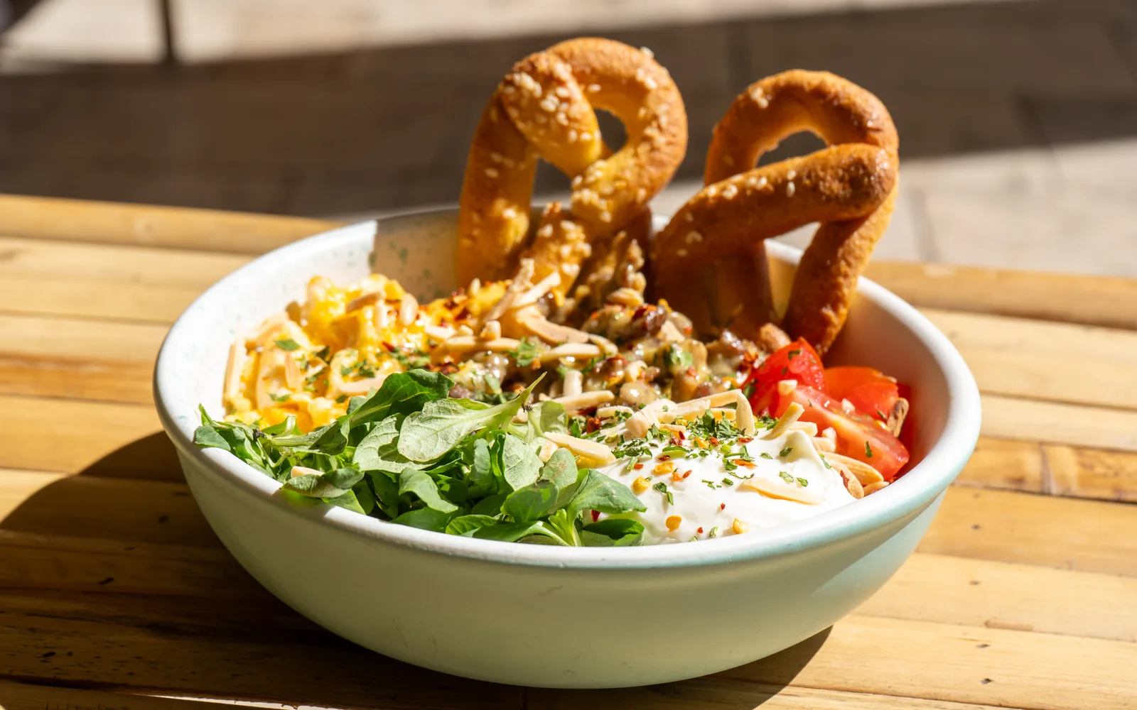 The Philly Cheese Steak Protein-Bowl seen low from the side at Cookie Beacon Budapest: two sesame keto pretzels standing upright behind scrambled eggs, sirloin in cheese sauce, lamb's lettuce, homemade sour cream and cherry tomatoes in a pale mint bowl