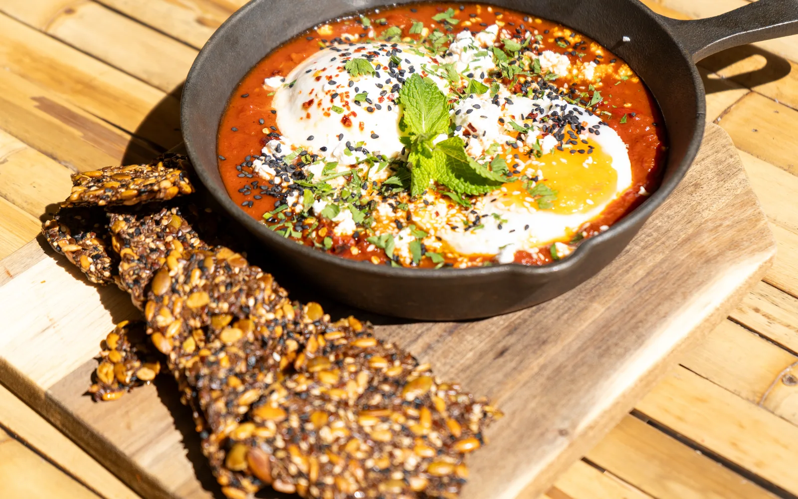 The Shakshuka Burrata on a sunny bamboo table at Cookie Beacon Budapest: the cast-iron skillet on a wooden board, with a stack of homemade seed crackers studded with whole seeds in the foreground