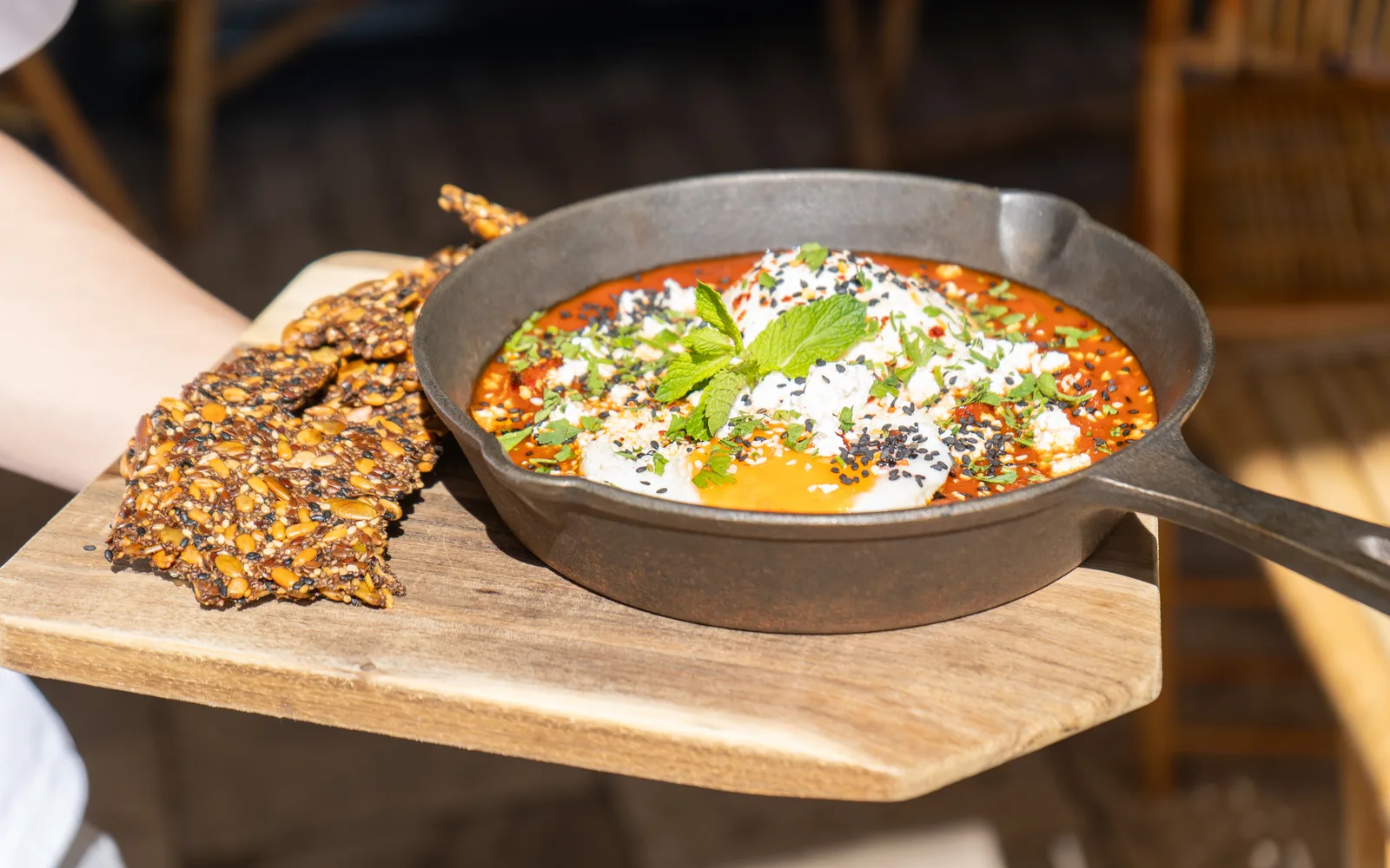 The Shakshuka Burrata carried out on a wooden board at Cookie Beacon Budapest: a cast-iron skillet of red tomato and pepper sauce with a sunny side up egg, a white dome of burrata, feta crumble, black and white sesame and a sprig of green herbs, with dark seed crackers beside the pan