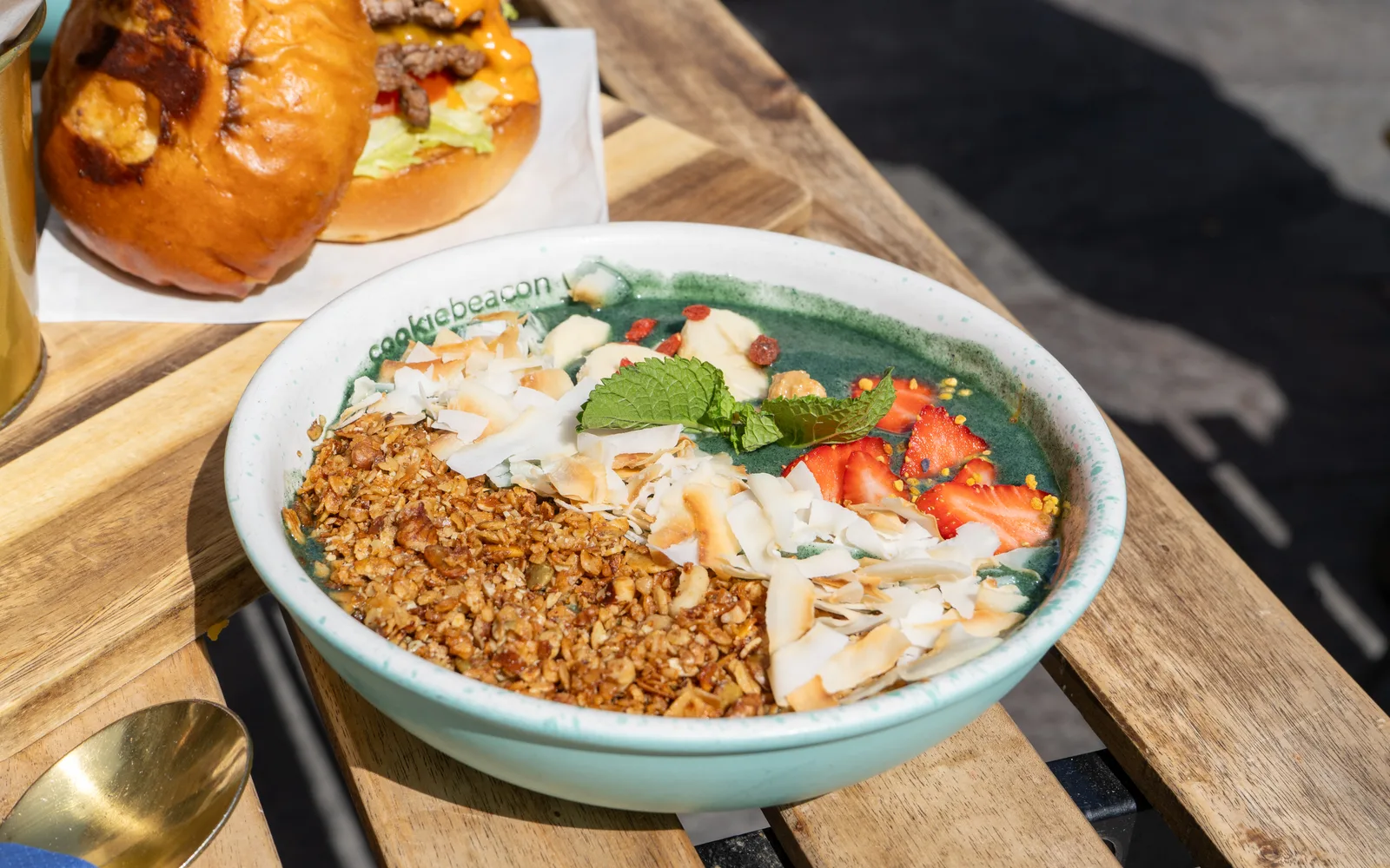 Spirulina Power Granola Bowl at Cookie Beacon Budapest: deep green coconut-milk base under honey-toasted granola, coconut flakes, banana, strawberries, goji and mint, on a wooden terrace table