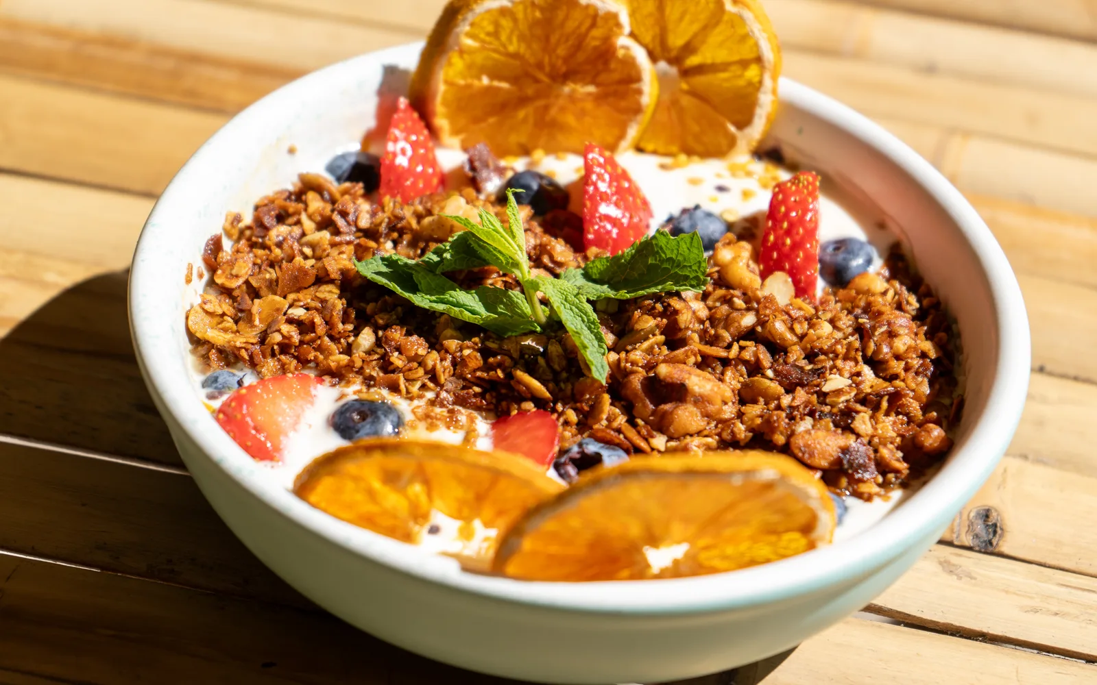 Close-up of the Yogurt & Honey Whipped Granola Bowl at Cookie Beacon Budapest: clusters of honey toasted granola with nut pieces and seeds, strawberries, blueberries, pollen granules and mint over whipped yogurt, with dehydrated orange slices at the top and bottom of the rim