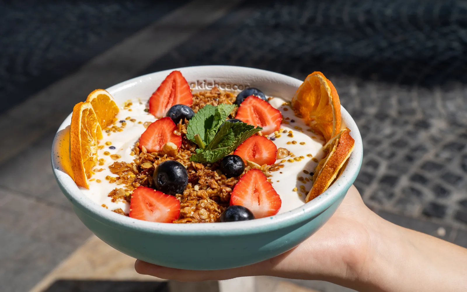 The Yogurt & Honey Whipped Granola Bowl held up in the sun outside Cookie Beacon Budapest: a pale blue bowl of white whipped yogurt topped with a heap of toasted granola, halved strawberries, blueberries, three slices of dehydrated orange at the rim, a sprig of mint and a scatter of yellow and orange pollen granules
