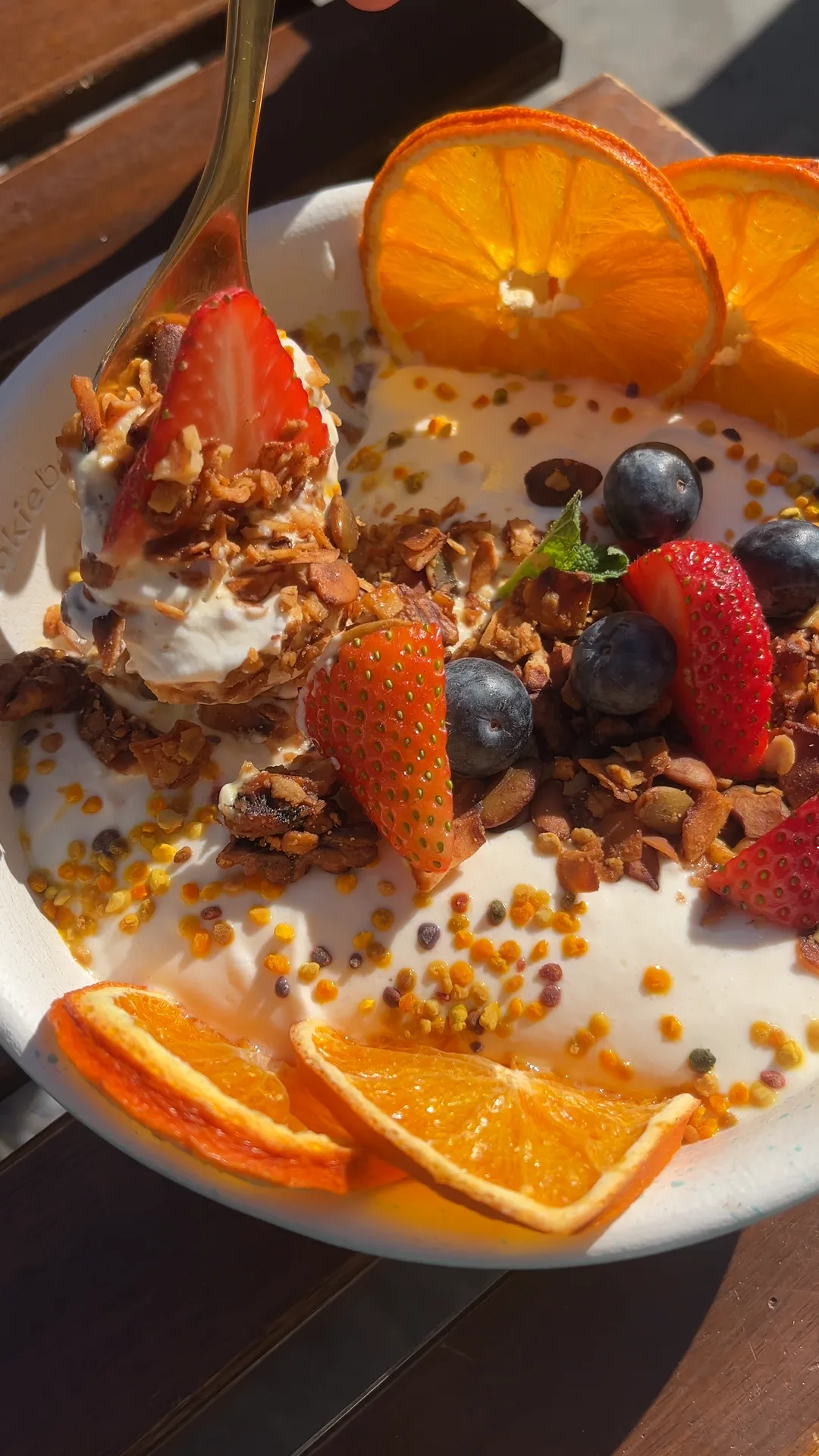 A gold spoon lifting whipped yogurt and granola out of the Yogurt & Honey Whipped Granola Bowl at Cookie Beacon Budapest, with strawberries, blueberries, pollen and dehydrated orange in the bowl