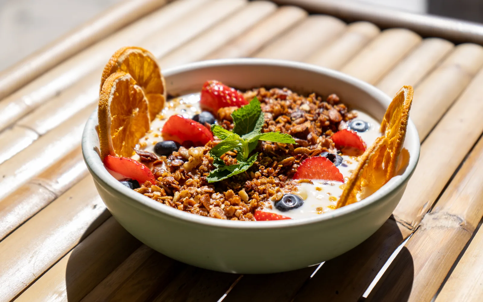 The Yogurt & Honey Whipped Granola Bowl on a bamboo table at Cookie Beacon Budapest: toasted granola over whipped yogurt, with strawberries, blueberries, dehydrated orange slices and mint on top